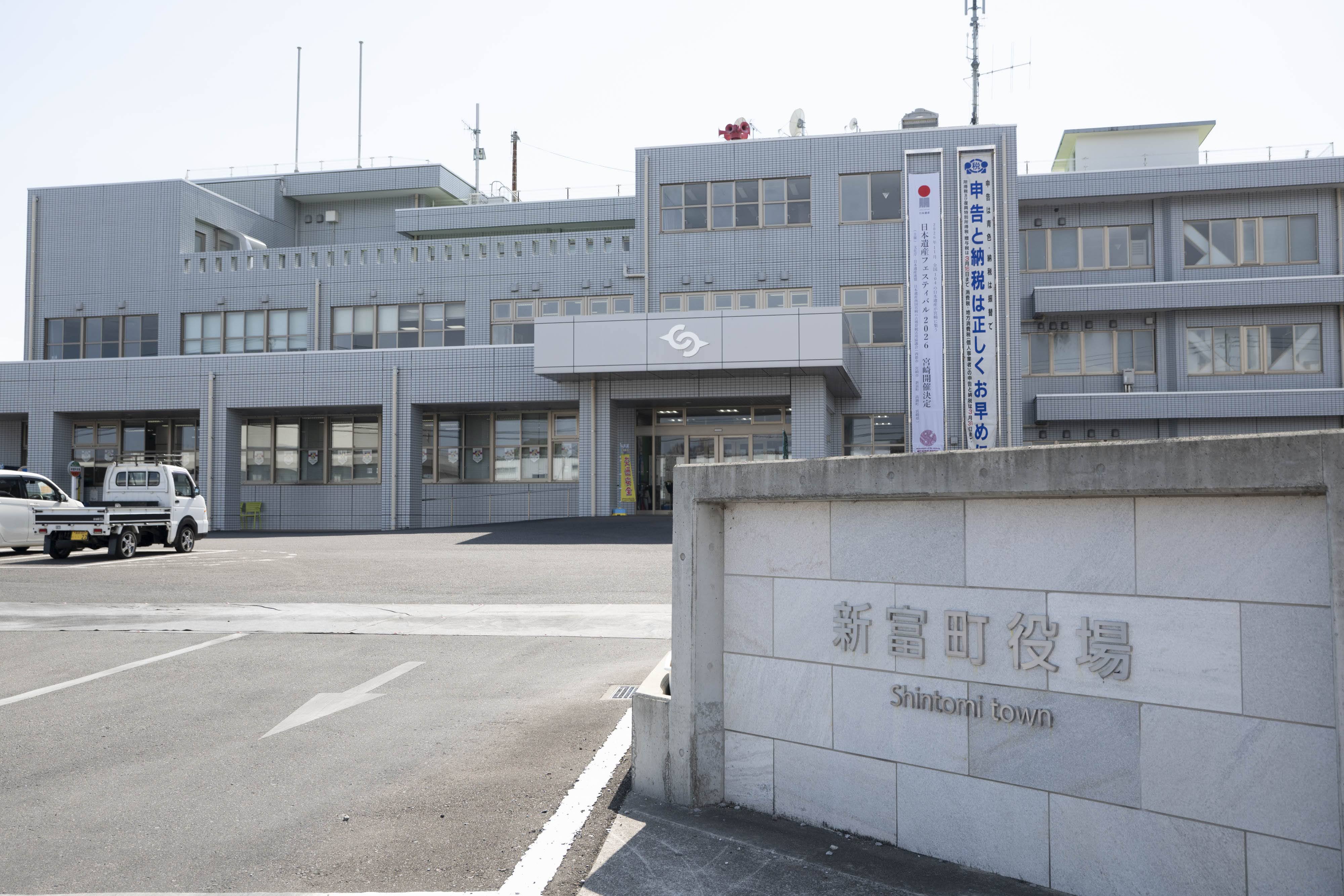 灰白色の庁舎が正面に映っている。手前右側に「新富町役場 Shintomi town」という字が写っている。庁舎正面には縦型の懸垂幕が掲げられ、庁舎のエントランス上部には町のシンボルマークが掲示されている。