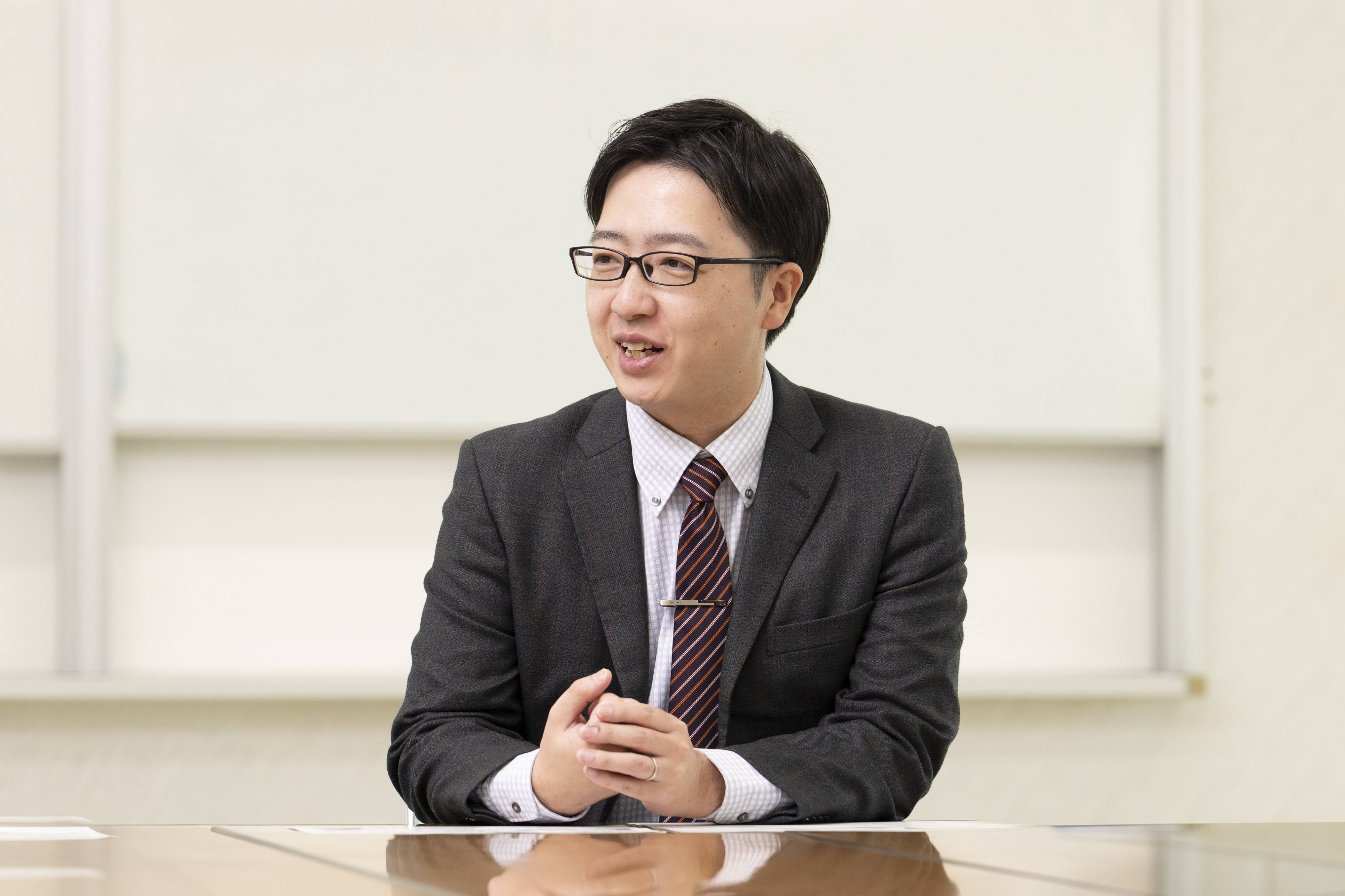 新富町総務課の吉野主査（※所属・職名などは取材時のもの）がテーブルに手を組んで着席しながら話している様子。
