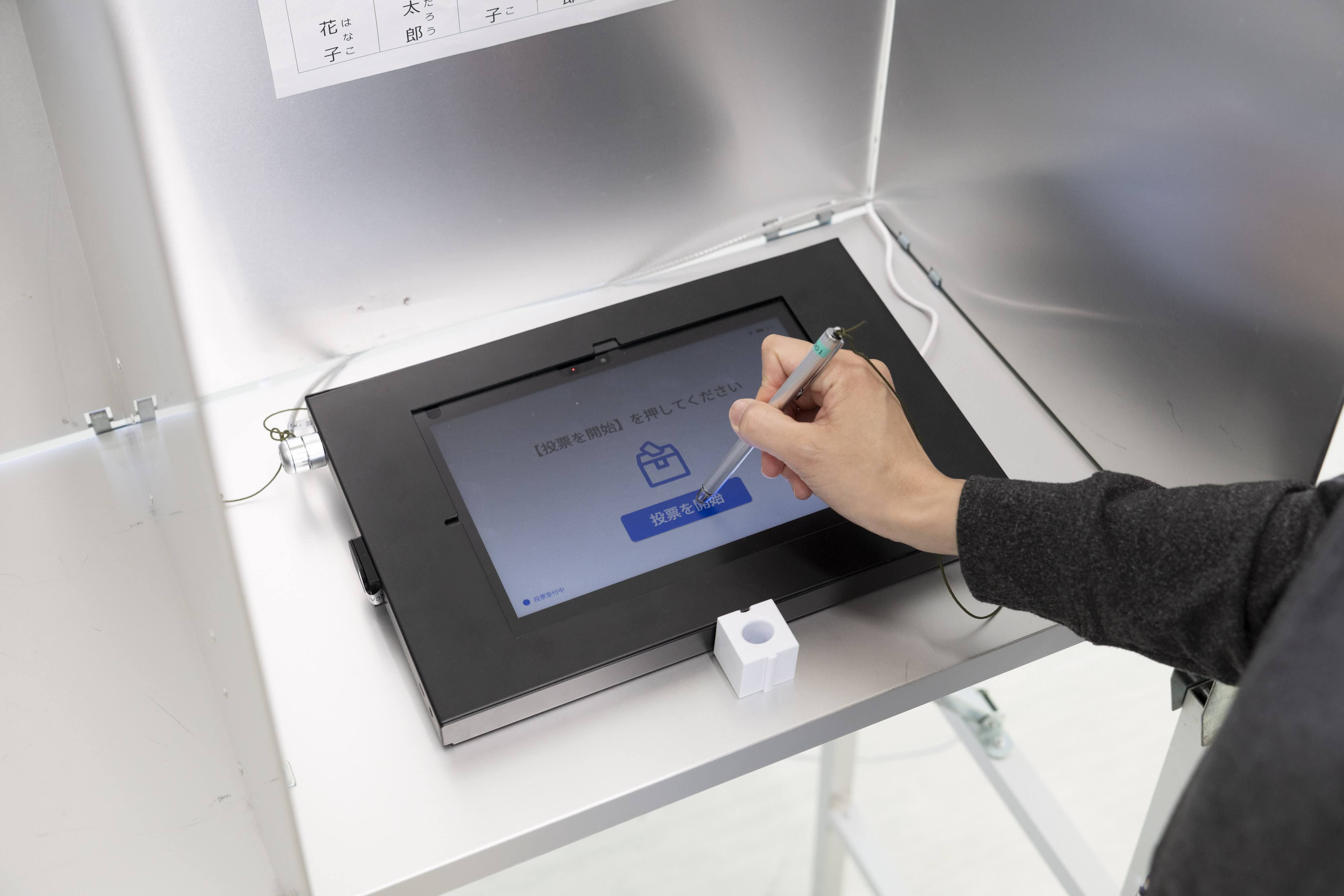 仕切りで囲まれた記載台に、黒いフレームのタブレット型電子投票端末が設置されている。画面には「【投票を開始】を押してください」のメッセージと投票箱のアイコン、青いボタン「投票を開始」が表示されている。スタイラスペンを持ち、画面を操作しようとしている手元が写っている。端末の手前にはペン立てが置かれている。背景の上部には候補者名が書かれた掲示の一部が見える。