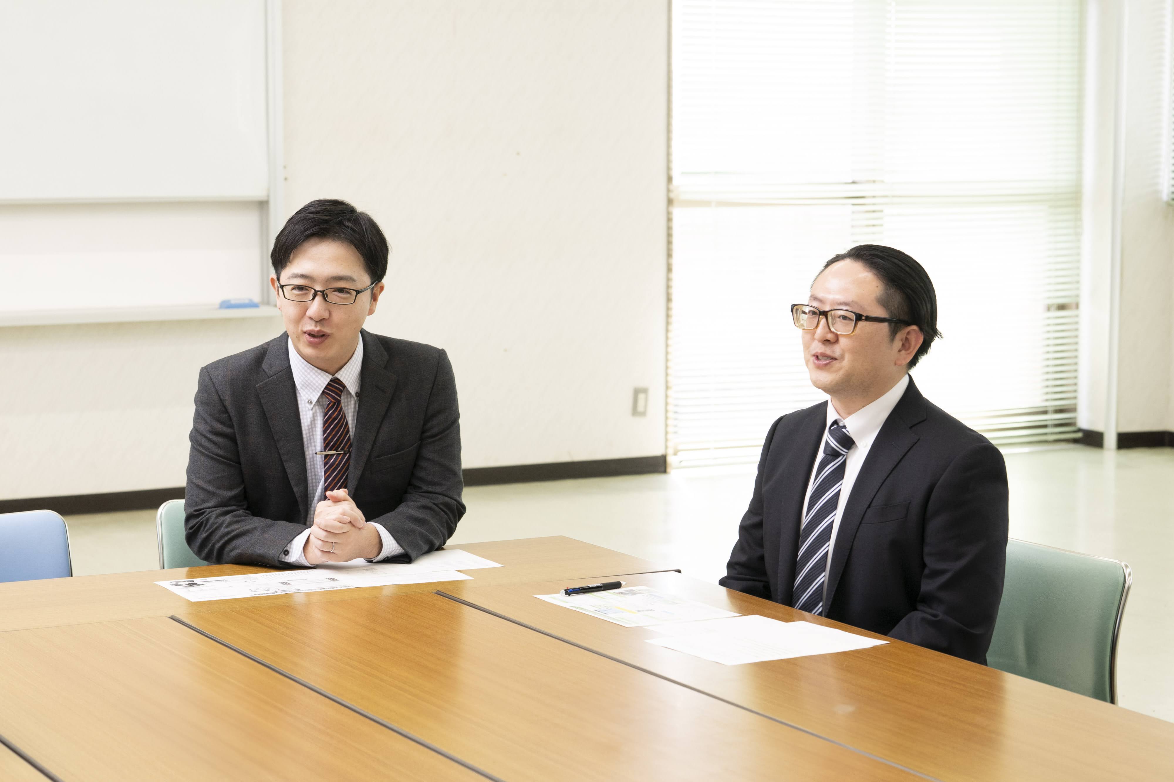 新富町総務課の吉野主査（左）と甲斐係長（右）が会議室の木目調テーブルに着席して話している様子。