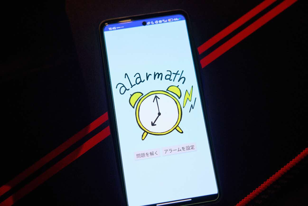 スマートフォンに表示されたアプリ「alarmath（アラーマス）」のトップ画面の写真。目覚まし時計のイラストと2つのボタンが表示されている。画面中央には手書き風のタッチで描かれた黄緑色の目覚まし時計のイラストと、右側に黄色い稲妻マークが描かれている。アプリ名「alarmath」は手書き風フォントで上部に表示されている。画面下部には「問題を解く」と「アラームを設定」の2つのボタンが横並びに配置されている。