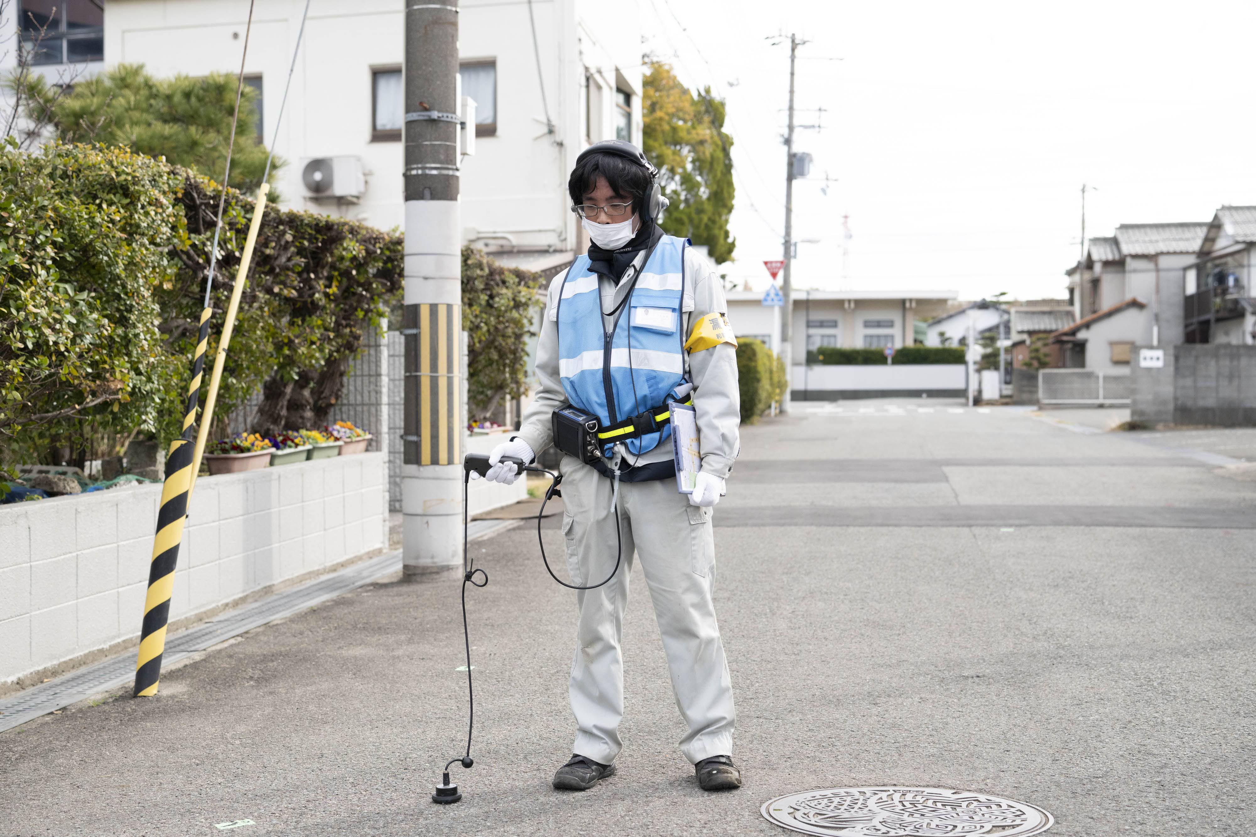 ヘッドセットを着用した作業服・青色ベスト姿の人物が、住宅街の道路上に立っている。路面音聴機を路面にあて、音聴調査をしている。脇には書類を抱えており、右手にはマンホールが見える。背景には一般住宅や電柱、植木が並ぶ住宅街の風景が広がっている。