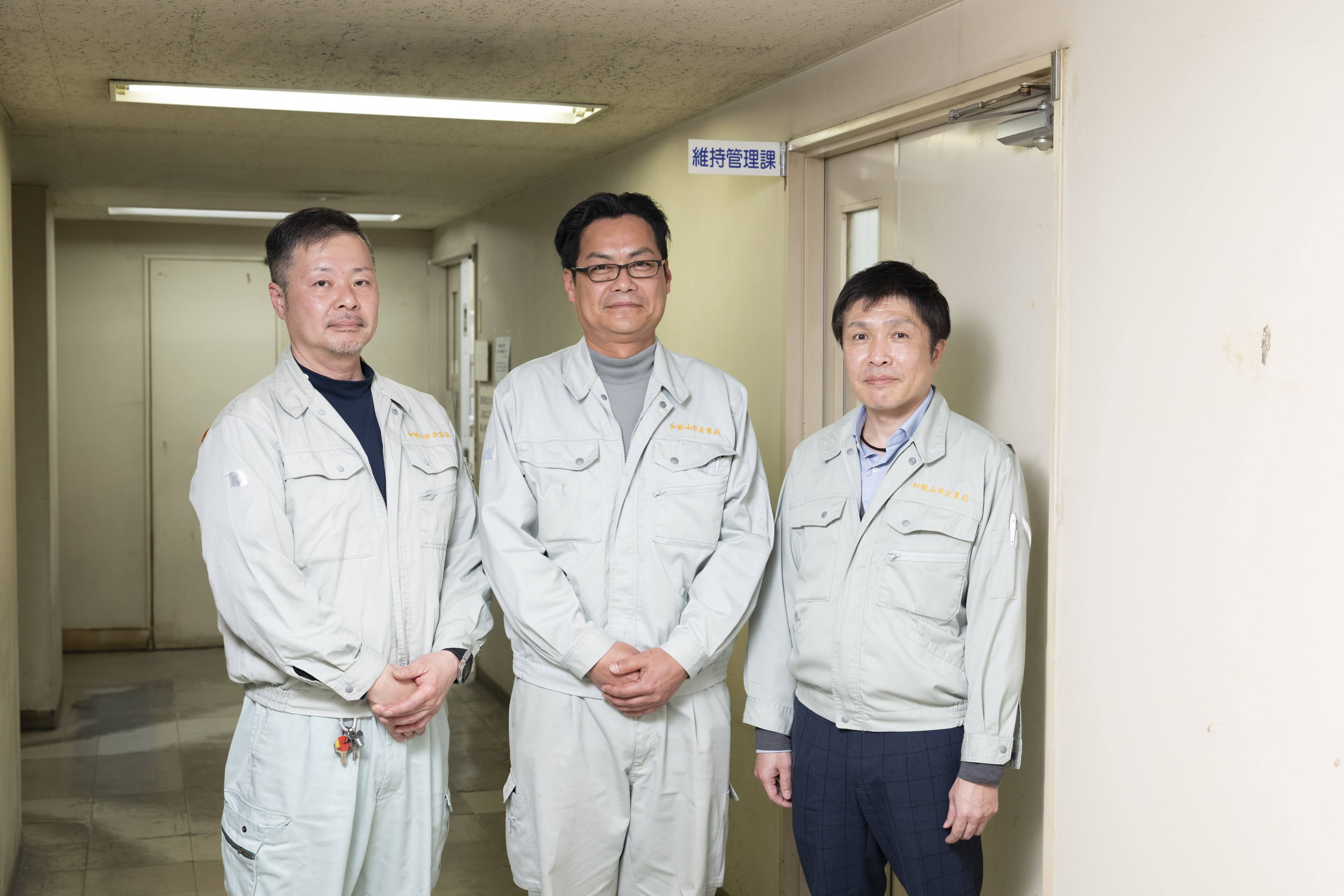 和歌山市企業局水道工務部維持管理課の職員が廊下で横一列に並んでいる様子を正面から撮影している。右側の扉には「維持管理課」と書かれたプレートが掲示されている。