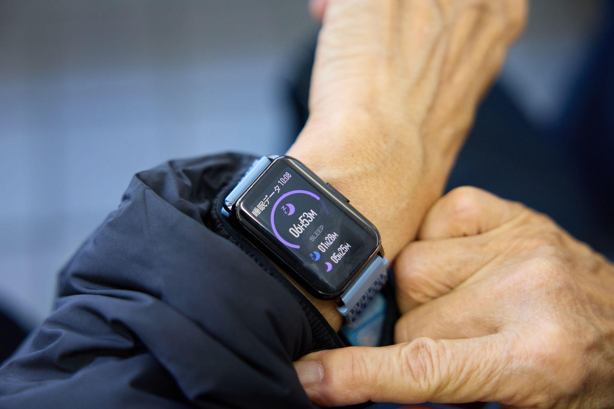 まちの保健室利用者の手首に装着されたスマートウォッチの睡眠データ画面。データ画面には「睡眠データ 10:08」と表示されている。紫色の円グラフとともに「06h53M」（総睡眠時間）、「SLEEP」、「01h28M」（浅い睡眠）、「05h25M」（深い睡眠）の数値が表示されている。