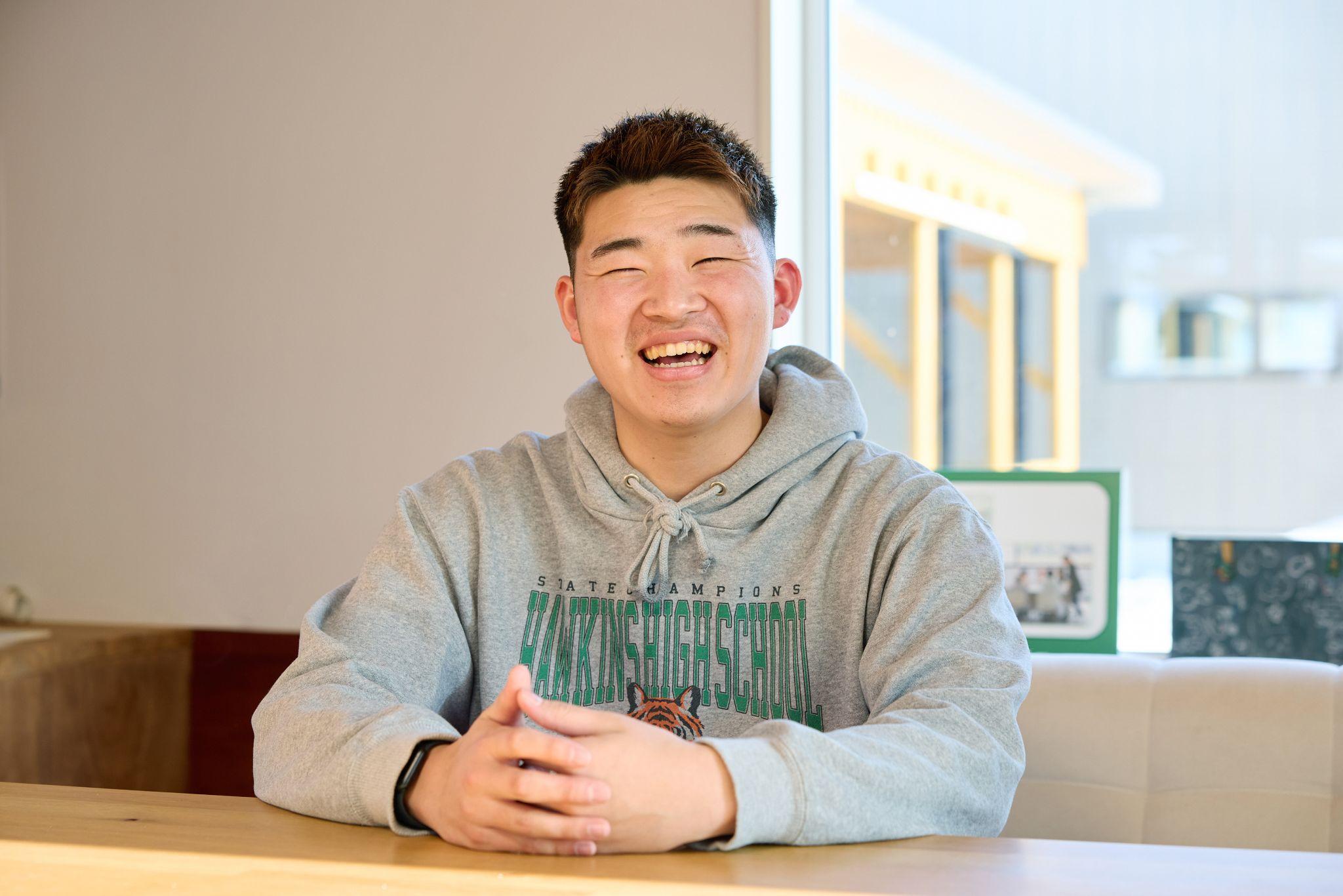 インタビューに応じる今村氏の写真。室内の木製テーブルに両腕を乗せて座っている。グレーのパーカー姿。背景には窓から外の建物が見える。