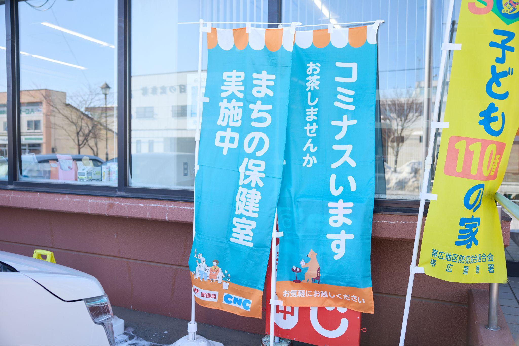 更別村郵便局の屋外の様子。2本の水色ののぼり旗が立てられており、左のぼりに「まちの保健室 実施中」、右のぼりに「コミナスいます お茶しませんか」と記載されている。
