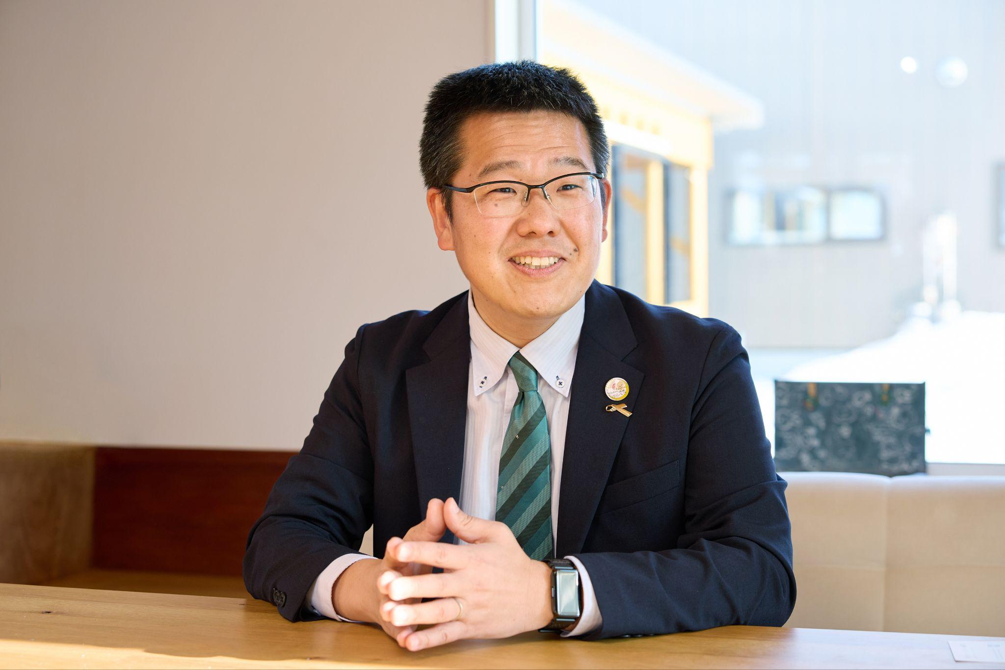 インタビューに応じる更別村の八木上席主査の写真。木製テーブルに両手を組んで座っている。紺色のスーツ、白いシャツ、グリーン系ストライプのネクタイを着用し、眼鏡をかけている。背景には窓から外の建物が見える。