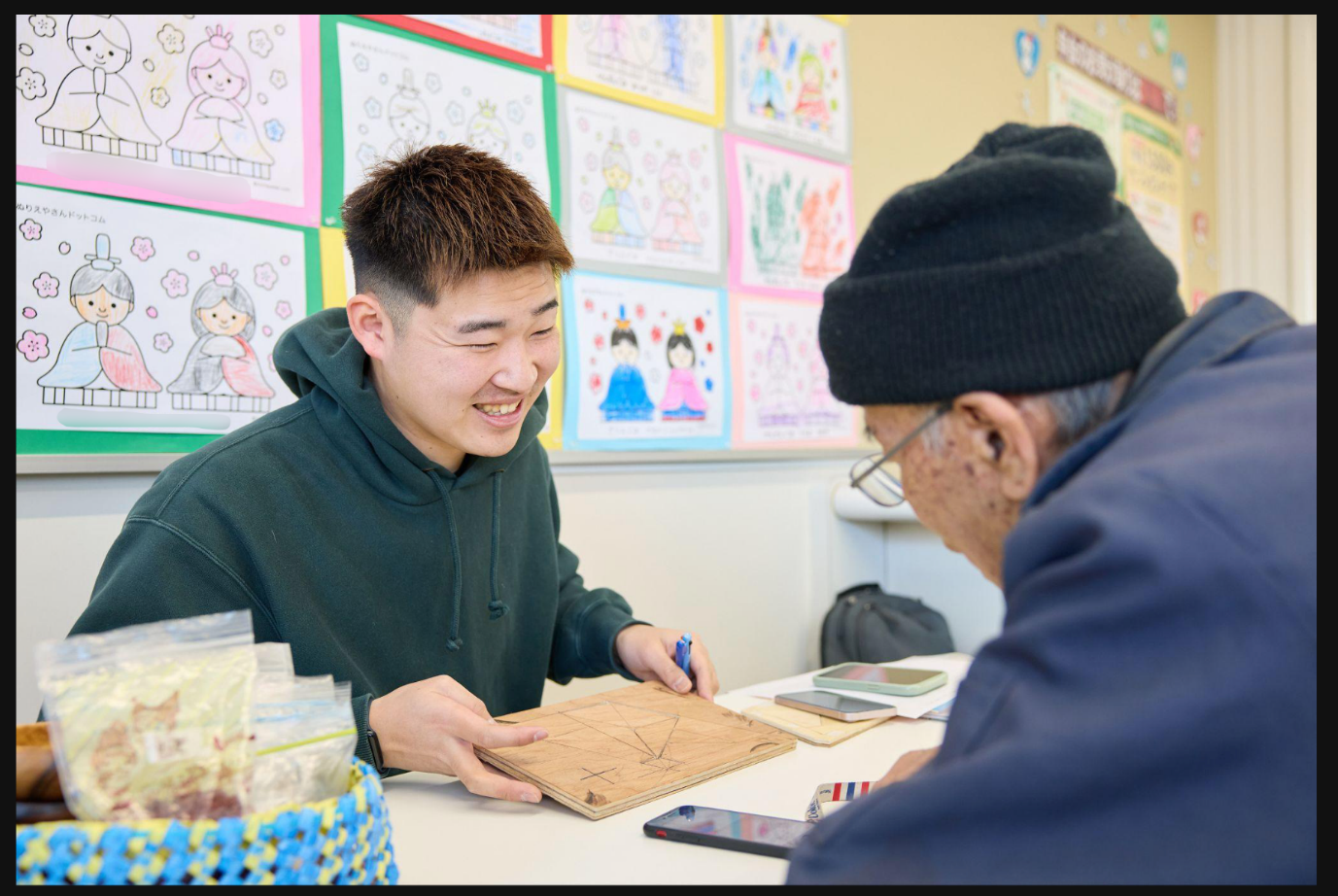 デジタル推進委員として活動するコミュニティナースの今村氏の写真。室内のテーブルを挟んで、緑のパーカーを着た今村氏と（左側）と、黒いニット帽・紺のジャケットを着た男性（右側・後ろ向き）が向かい合っている。テーブルの上にはスマートフォンや小袋に入ったお菓子、青いチェック柄のかごが置かれている。