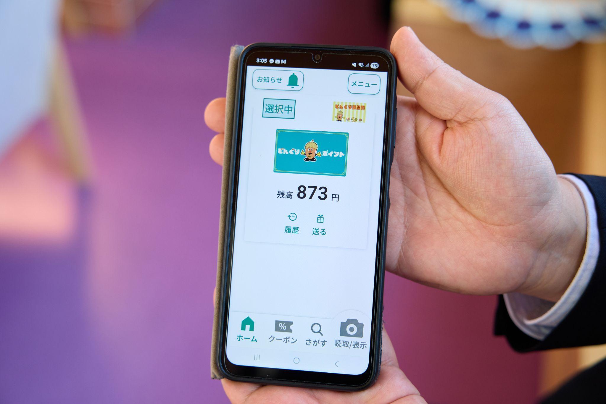 更別村の「デジタルどんぐりスタンプ」を映し出したスマートフォンの画面を撮影した写真。八木氏がスマートフォンを手に持ち、「どんぐりポイント」アプリのホーム画面を表示している。画面には「どんぐりポイント」アプリが表示されており、残高873円と記載されている。画面上部には「お知らせ」「メニュー」ボタン、「選択中」の表示があり、下部のナビゲーションバーには「ホーム」「クーポン」「さがす」「読取/表示」の4つのタブが並んでいる。