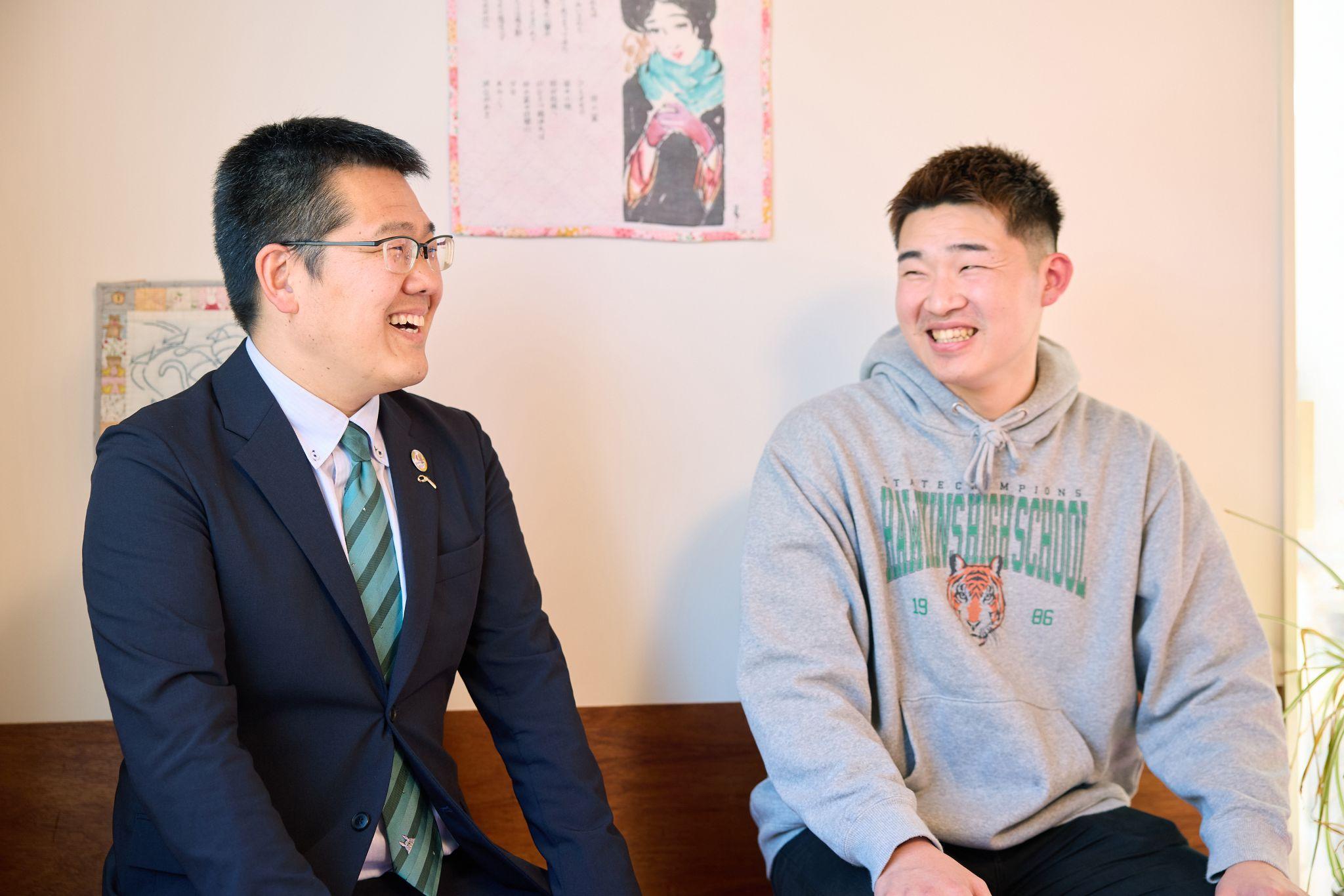 インタビューに応じる八木氏と今村氏が並んで座って対話している写真。左側に座る八木氏はスーツにストライプのネクタイ、眼鏡を着用。右側に座る今村氏に向かって笑顔を向けている。今村氏はグレーのパーカーを着用し、笑顔を見せている。