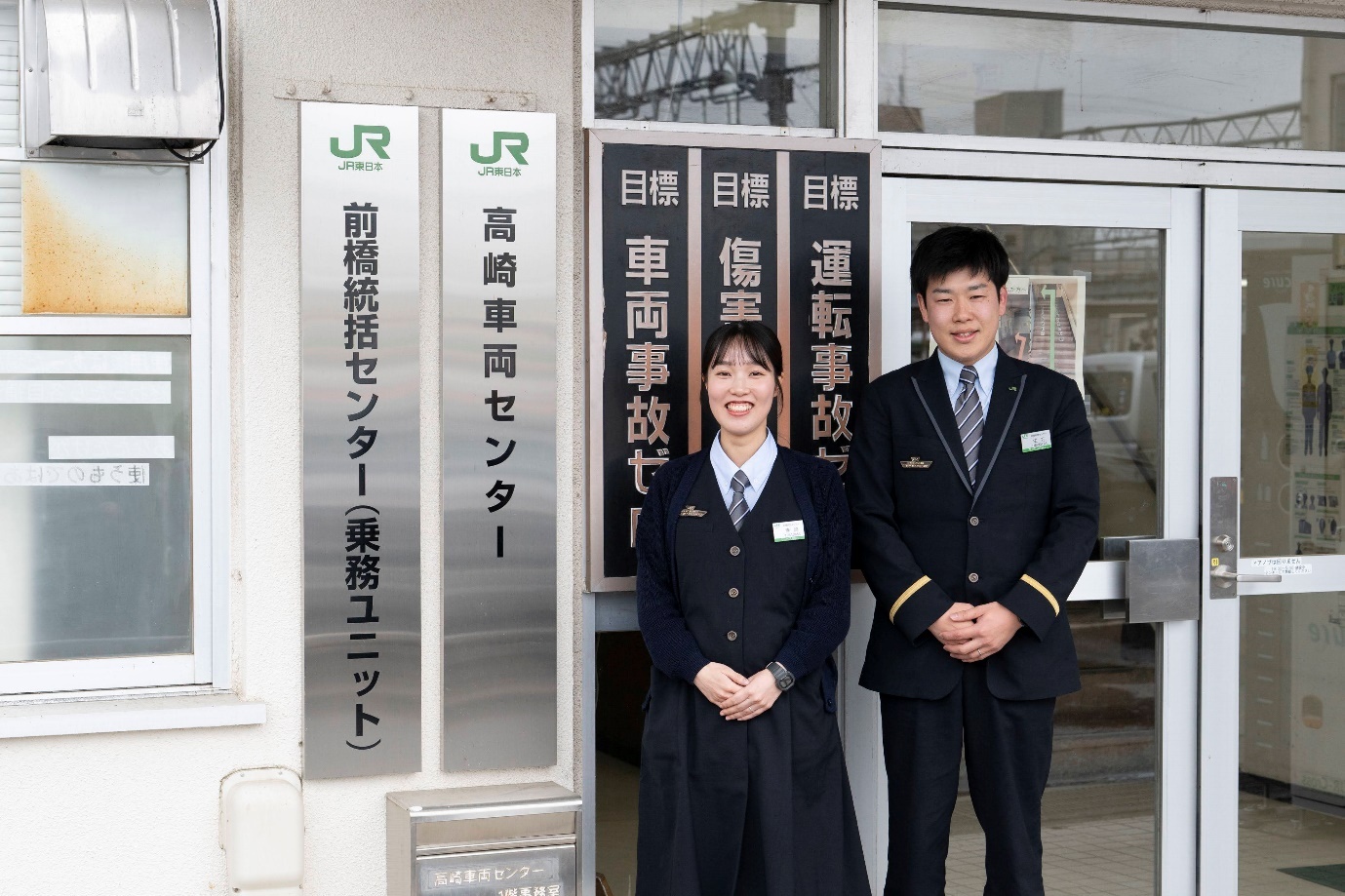 JR東日本の制服を着用した前橋統括センター乗務ユニットの寺嶋彩乃氏（左）と生方聡氏（右）が建物入口の前で並んで立っている。背後には「JR東日本 高崎車両センター」「前橋統括センター（乗務ユニット）」の表示板と、「目標 車両事故ゼロ」「目標 運転事故ゼロ」などと書かれた安全目標の掲示板が見える。