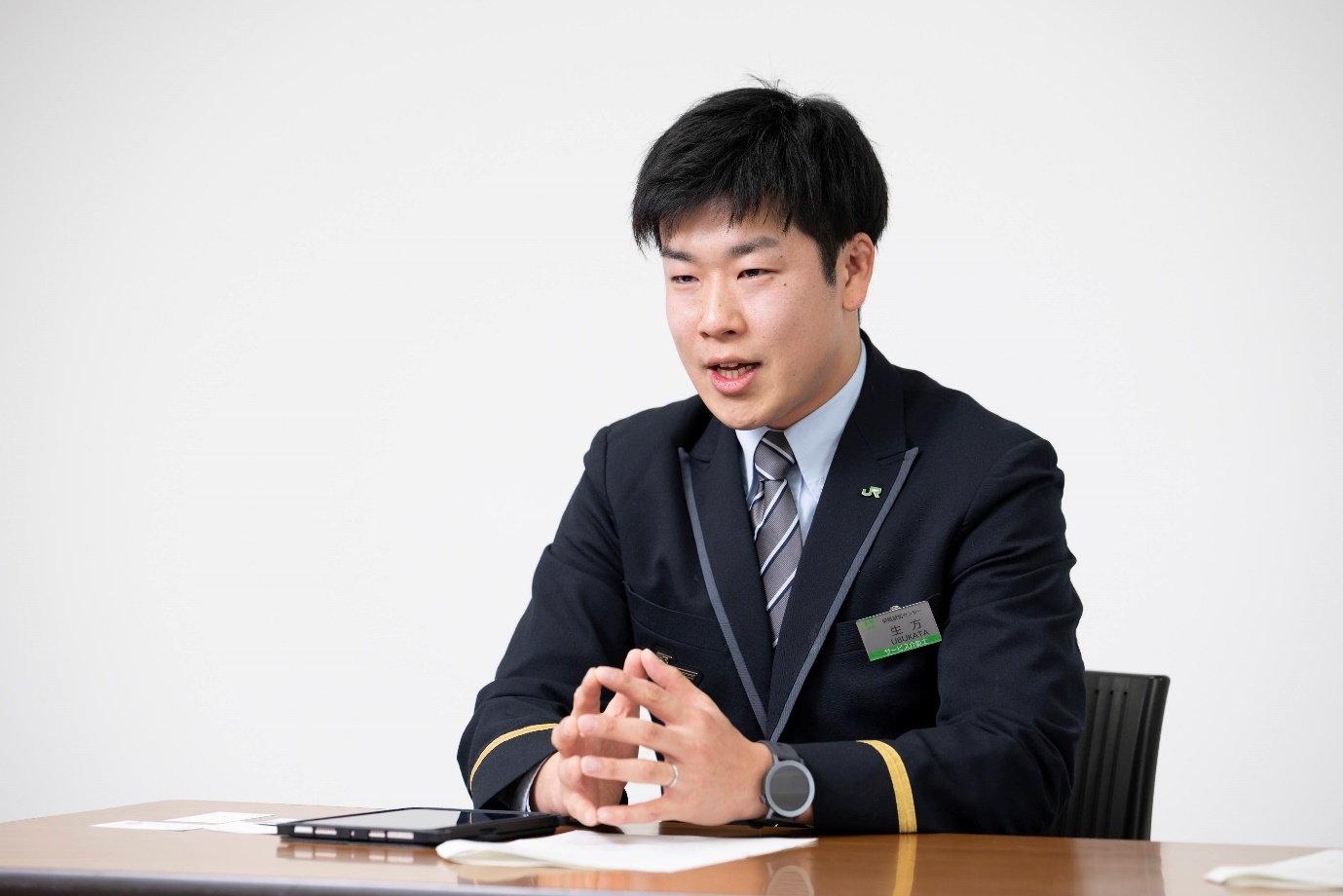 インタビューに応じる生方聡氏。JR東日本の制服（袖に金のライン入り）を着用し、白い壁紙を背景にテーブルの前に座っている。胸にはネームプレートをつけており、JRのバッジも見える。