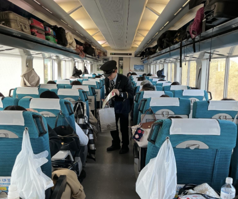 特急草津・四万の車両内で実施した「デジタルよろず相談所」の様子。青緑色のシート（白いヘッドレスト付き）が並ぶ列車の車内で、制服を着用したJR東日本の職員が通路を歩き、相談対応にあたっている。座席は概ね乗客で埋まっており、荷物棚には多数のバッグやスーツケースが置かれている。
