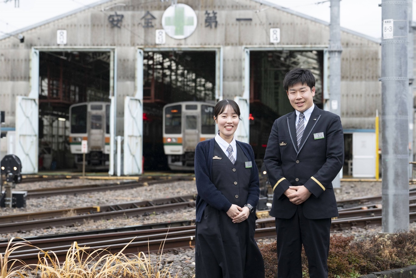 JR東日本の制服を着用した前橋統括センター乗務ユニットの寺嶋彩乃氏（左）と生方聡氏（右）が、高崎車両センターの車両格納庫前を背景に立っている。背後の格納庫には「安全第一」の文字と緑十字のマークが掲げられており、格納庫内にはJR東日本の列車が停車している。