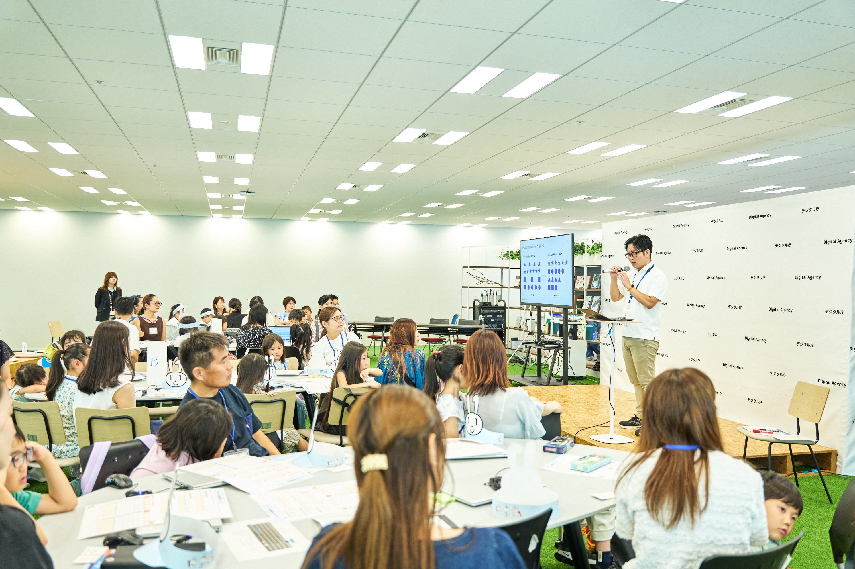 デジタル庁(Digital Agency)のロゴが繰り返し印刷されたバックパネルを背景に、デジタル庁職員がマイクを持ってステージに登壇している。会場には大型モニターが設置されており、丸や星などの図形が示されたスライドが映し出されている。会場内には大人と子どもが混在する多数の参加者がテーブルに着席し、資料やノートパソコンを手元に置いて講演を聴いている。床には人工芝が敷かれている。