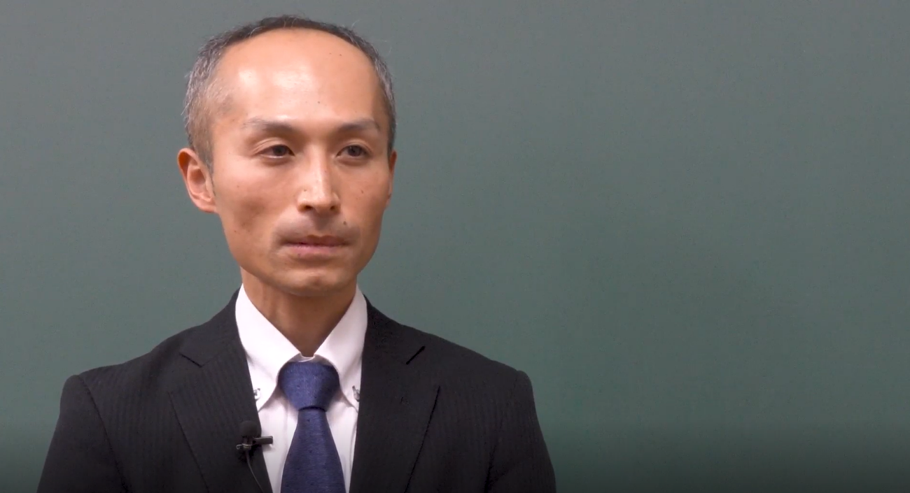 インタビューに応じる太田教諭。黒板の前でネイビーのネクタイを着用したスーツ姿でインタビューに応じている。