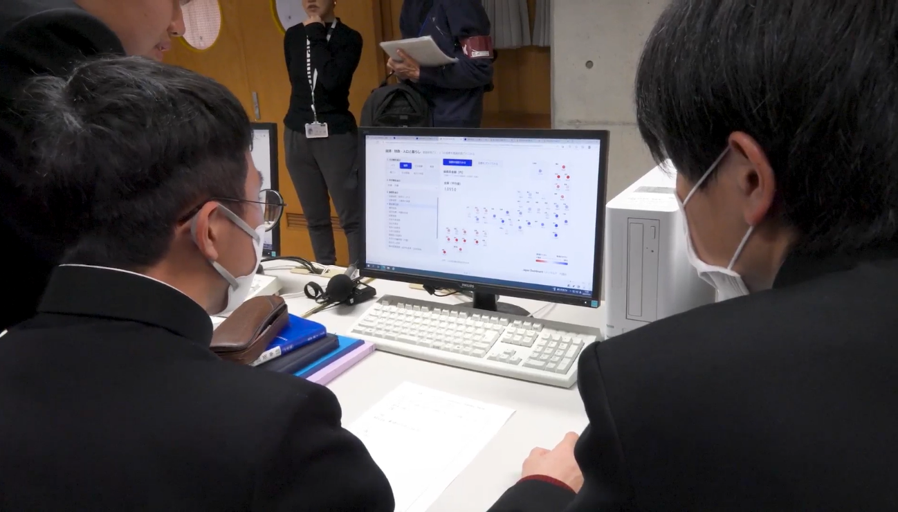 室内で、マスクを着用した黒い詰襟の学生服を着た高校生2名が、デスクに置かれたデスクトップPCのモニターを並んで覗き込んでいる。モニターには「居住・労働・人口と暮らし」に関するダッシュボードが表示されている。デスク上にはキーボード・書類・バインダーが置かれている。背景には別の人物がタブレットを手に立っている。