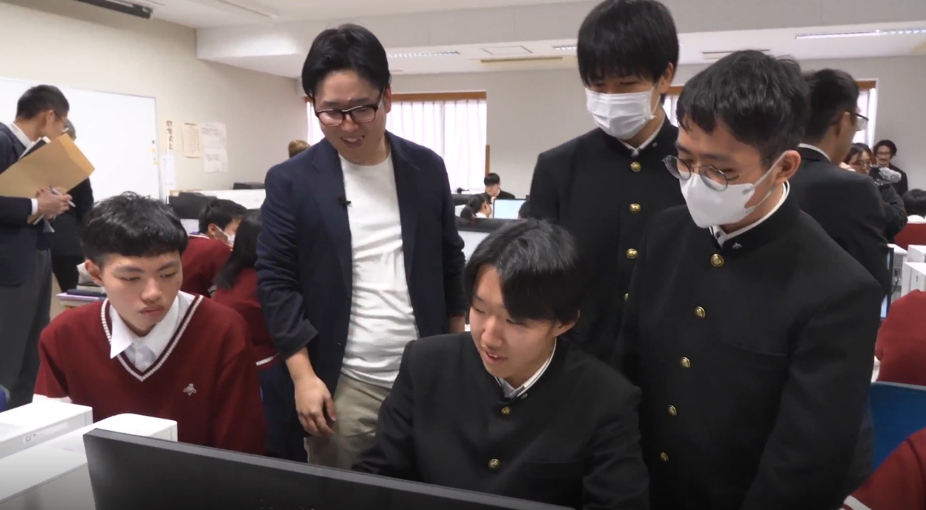 学校のコンピュータ教室で、詰め襟の学生服を着た男子生徒3名、えんじ色のセーターを着た男子学生1名、紺のジャケットに黒縁の眼鏡姿のデジタル庁職員がデスクトップパソコンを囲みつつ、画面を見ながら話し合っている。