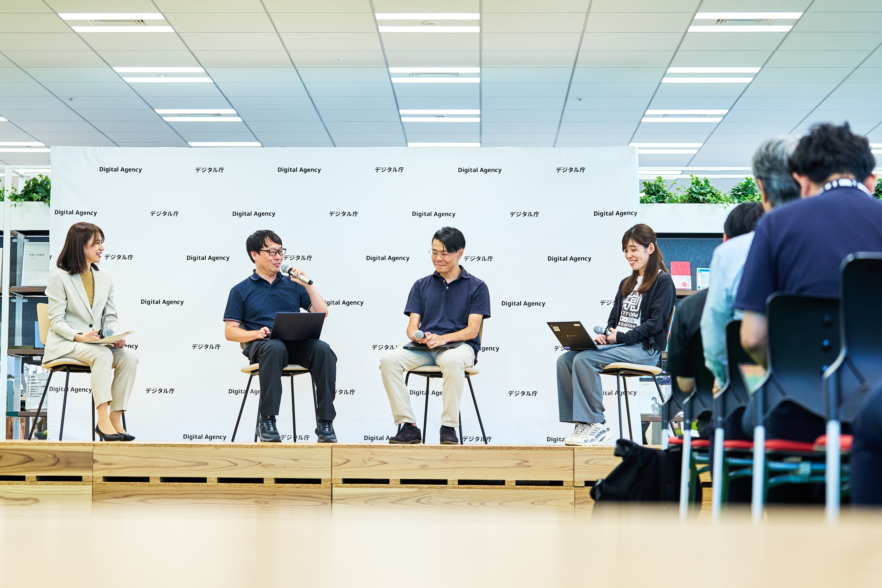 デジタルマーケットプレイス活用交流会に登壇したデジタル庁の柴原、吉田、グラファーの柏野氏、浜松市の内山氏が意見交換する様子を、正面から撮影した。ステージ上に横並びに置かれた椅子に着席し、左から2番目に座る吉田がマイクを持って発言している。ステージ背後には「デジタル庁」ロゴが入ったバックパネルが置かれている。登壇者がいるステージの手前には椅子が置かれ、参加者が着席して話に耳を傾けている。