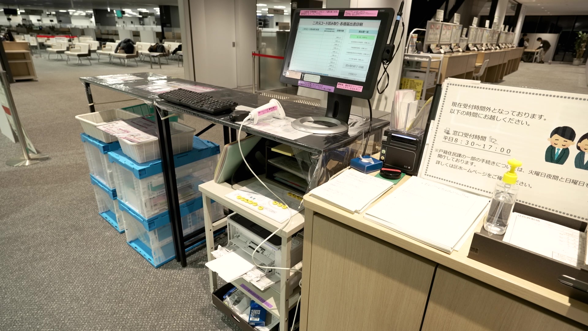 MKシステムを用いる窓口の様子。「二次元コード読み取り・各種届出書印刷」の画面を表示したモニター、バーコードスキャナー、キーボード、複数の青いコンテナボックスが置かれた作業台が写っている。