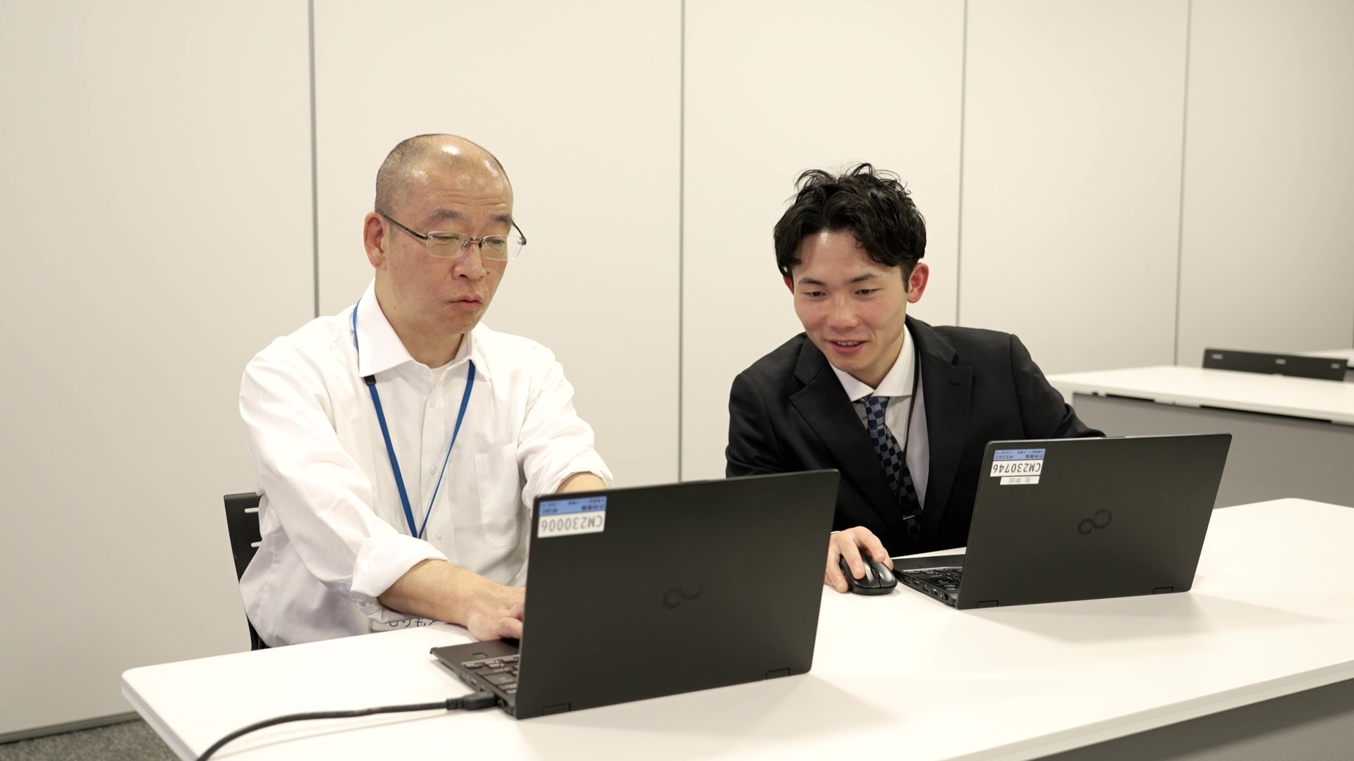 中野区デジタル政策課の森本主査（左）と戸籍住民課の岸田主査が白い長机に並んで着席し、各自の黒いノートPCを操作している様子。