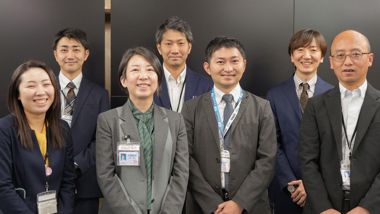 写真：熊本市 地域活動推進課・上野由起主査（前列左端）、井出祐輝主査（前列中央右）、防災計画課・大住浩二課長（前列右端）、甲斐怜志主査（後列中央）、西川智大主任主事（後列左）、情報政策課・北添友子副課長（前列中央左）、松永憲生主幹（後列右）