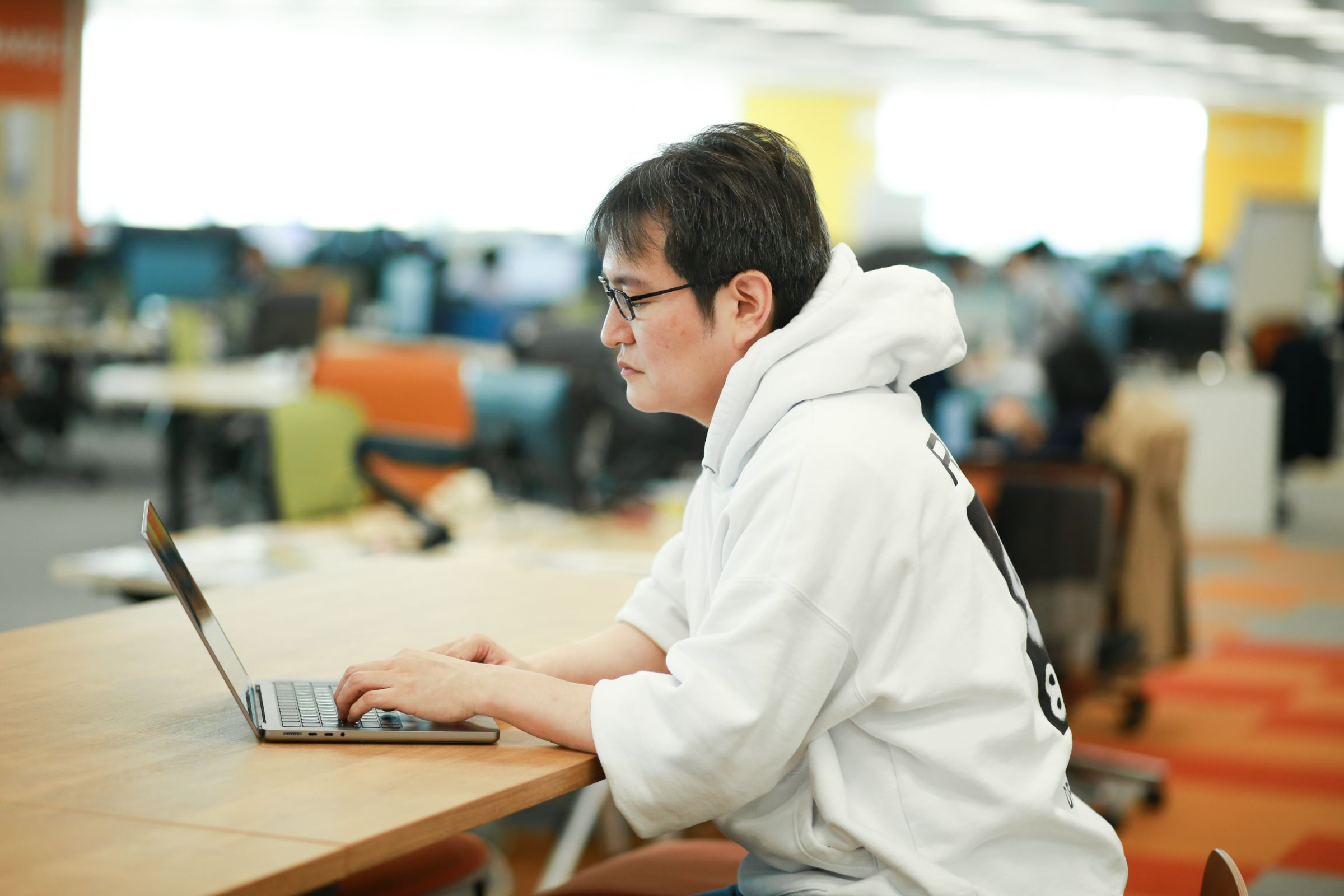 CTO 藤本真樹のウェストショット。オフィスのカウンターを前に座って、ノートパソコンを操作している様子を左側から撮影している。藤本は白いパーカーを着ている。