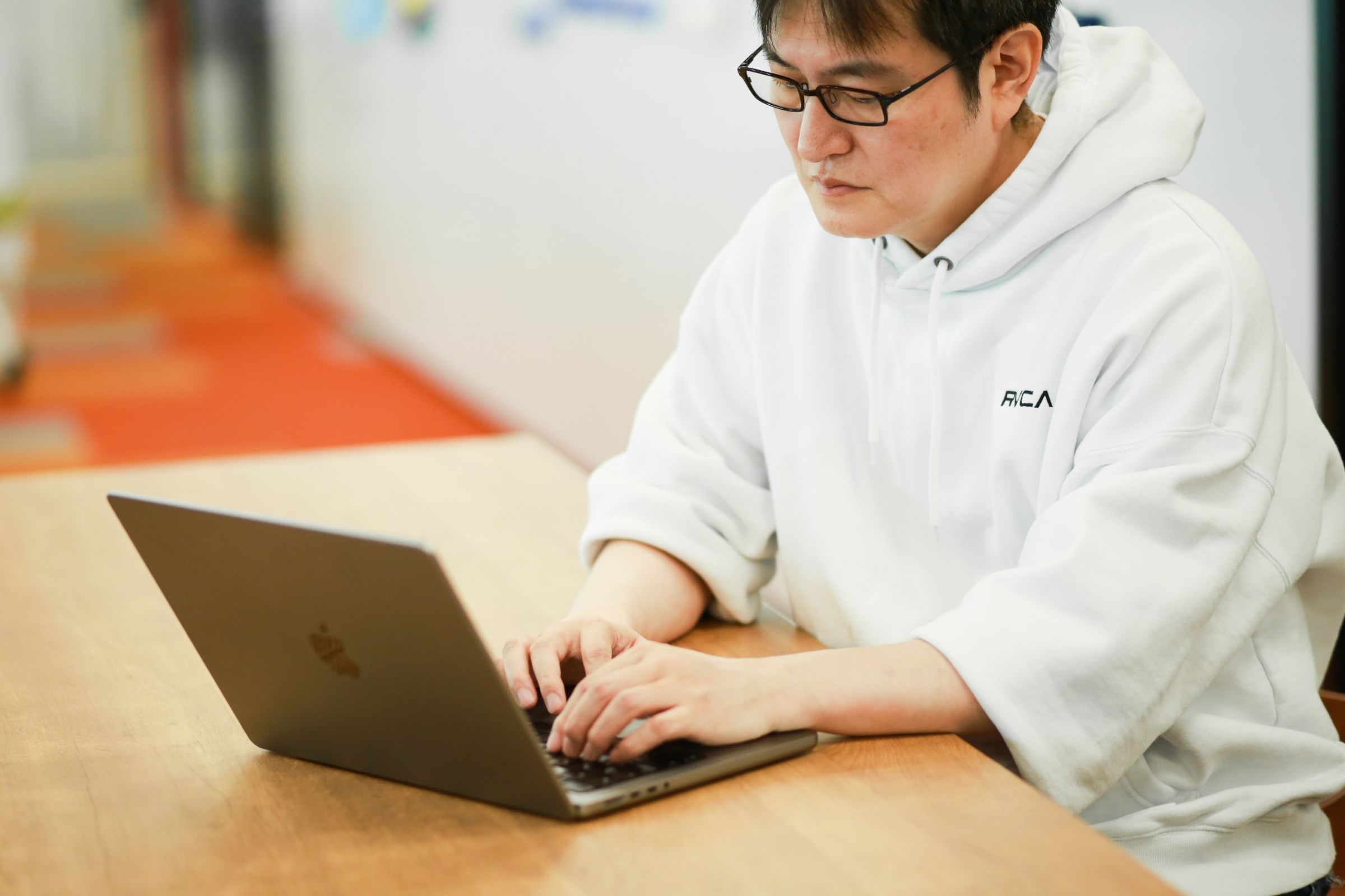 藤本のウェストショット。オフィスのカウンターを前に座って、ノートパソコンを操作している様子を左斜め上から撮影している。