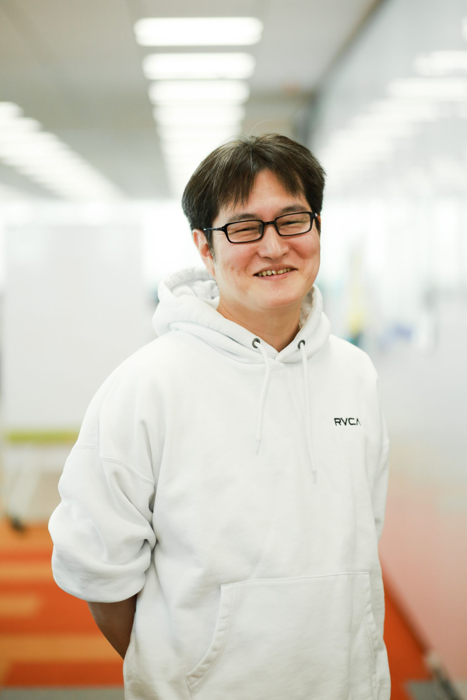 藤本のウェストショット。オフィスの中で、正面を向いてにこやかな表情で立っている。