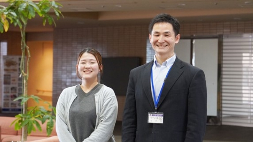 レンガ調の壁と観葉植物が見える明るい施設のロビーで、2名が並んで正面を向いている。 左側はグレーのカーディガンを着た若い女性(林氏)で、穏やかな笑顔を見せている。右側は黒いスーツに白いシャツを着た男性(武田氏)で、青いストラップの名札(IDカード)を首から下げており、笑顔を見せている。背景には掲示物やソファが確認できる。