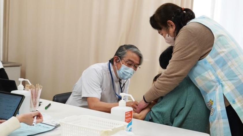 白衣とマスクを着けた白髪で高齢の男性医師が、診察台に座る子どもの腕(手首付近)を診察している。子どもの隣には水色のチェック柄エプロンを着けた女性(保護者)が付き添い、子どもを支えている。机の上には消毒液やトレーなどの医療用品が置かれており、白いカーテンを背景とした診察室の様子が映っている。