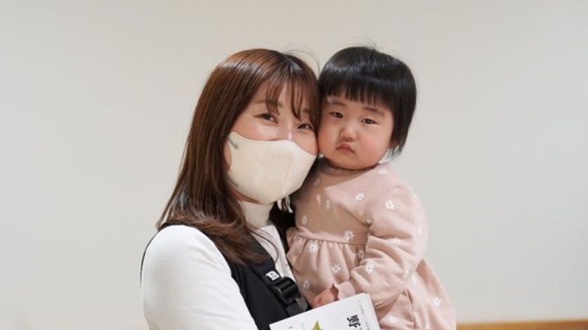 ピンク色の花柄の服を着た幼児(1〜2歳程度)を白いマスクを着けた白いタートルネック姿の若い女性(保護者)が正面に向けて抱いている。幼児は女性の頬に顔を寄せている。背景は白い壁。