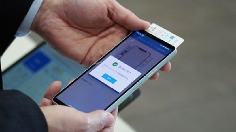 スーツ姿の男性の両手がスマートフォンを持ち、マイナンバーカードをスマートフォンで読み取る操作している。画面には緑色のチェックマークとともにマイ「読み取り完了」という完了メッセージが表示されている。スマートフォンの背面にはカードが当てられている。