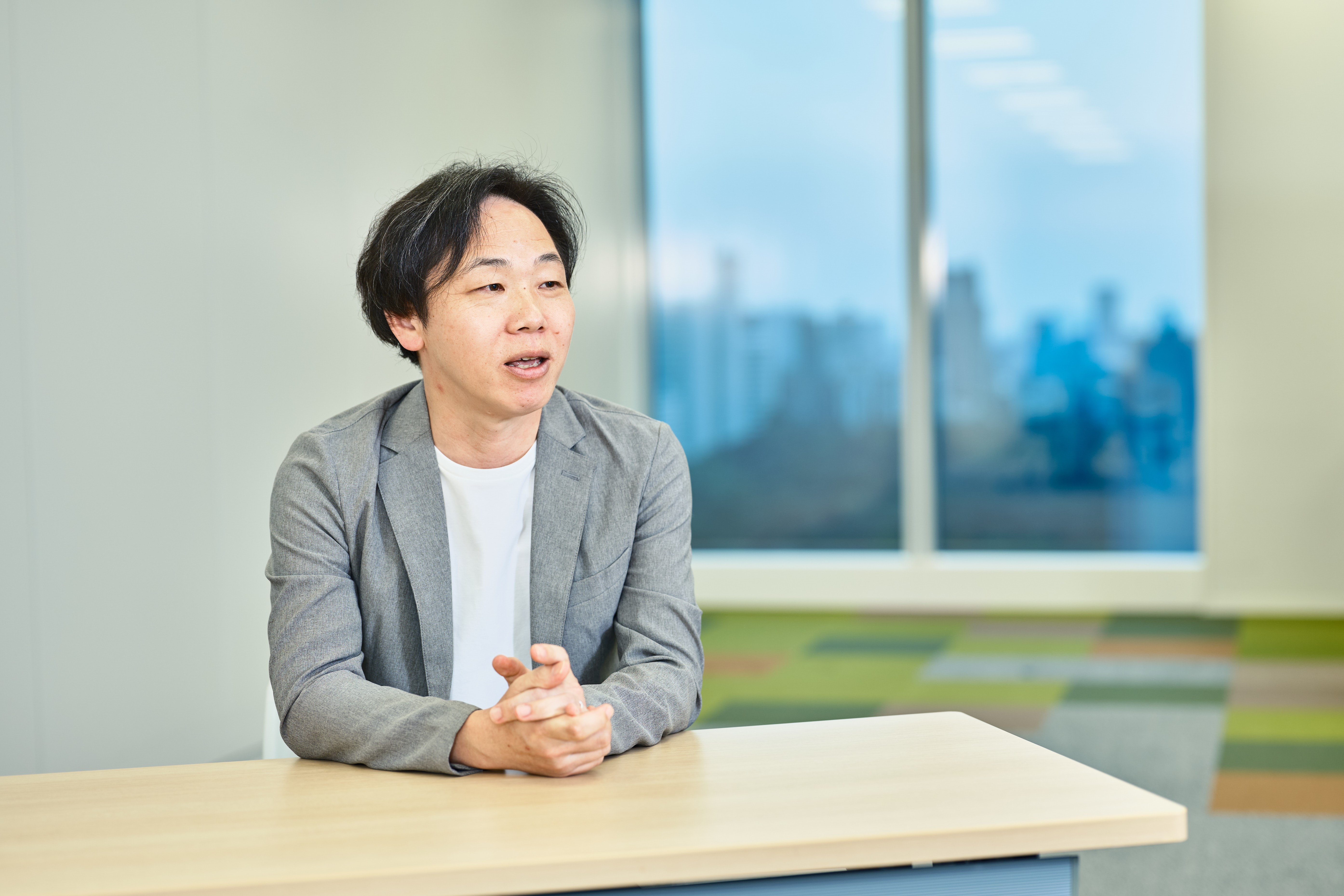 プロジェクトマネージャーの髙橋が、会議室と窓を背景に着席して話す様子を正面から撮影した写真。