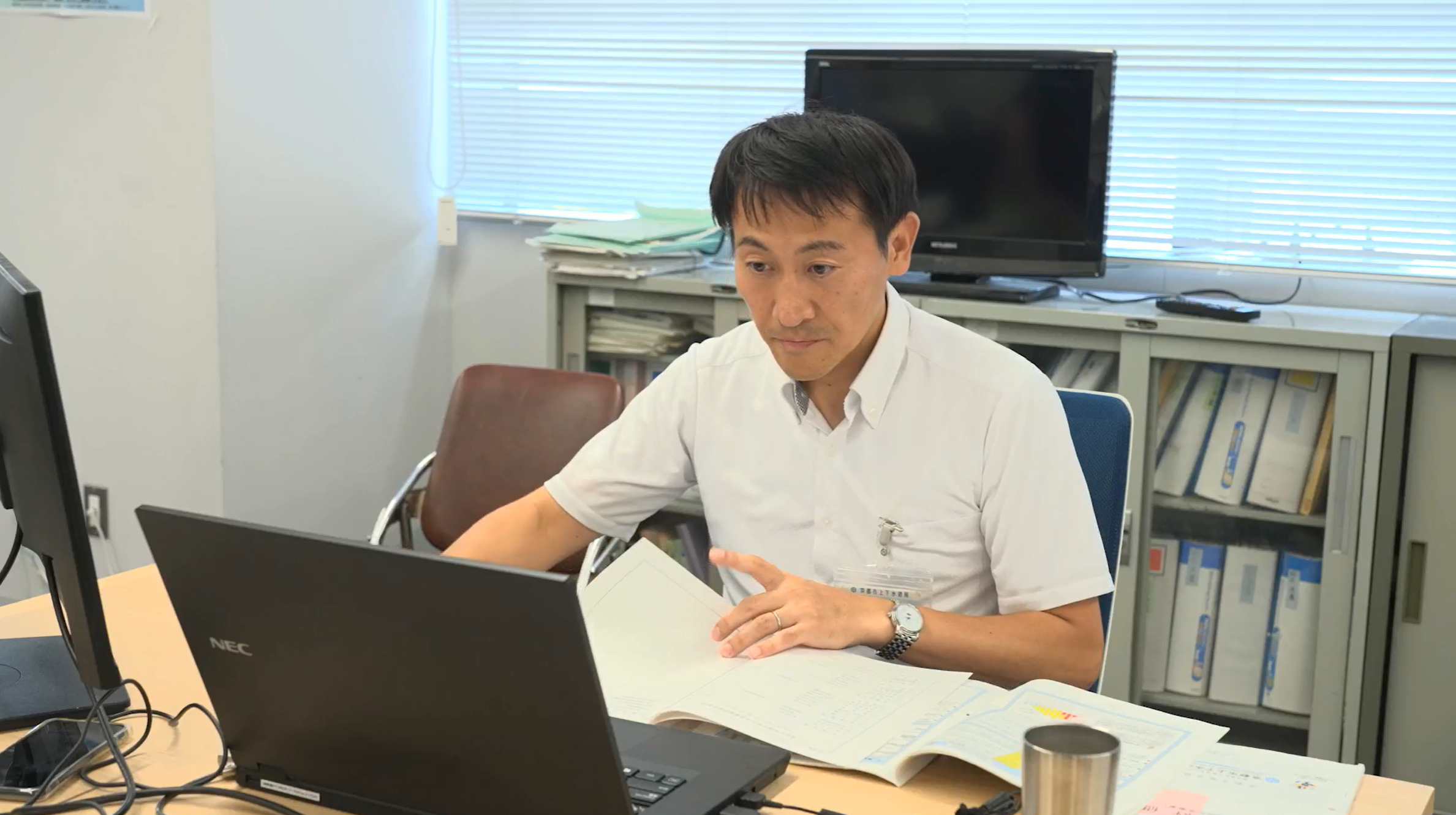 動画の切り取り画像。京都市上下水道局経営戦略室経営企画課の小堀善光課長がパソコンに向かって仕事をしている様子。手元には紙の資料も見える。