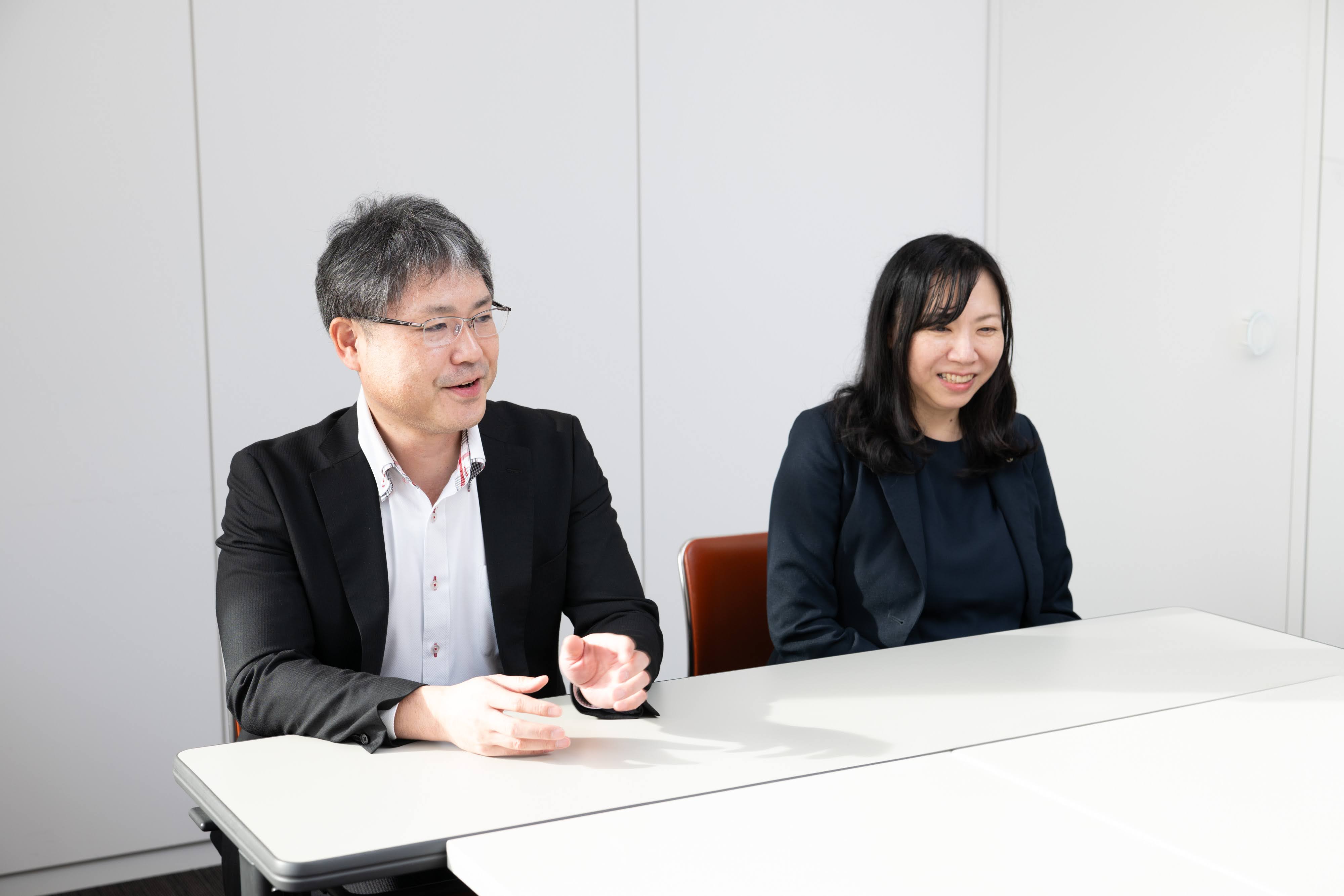 秋田市デジタル化推進本部の篠田副参事（左）と大倉主査（右）がインタビューに応じる様子。会議室の壁を背景に机に着席して話している模様を、正面向かって左手から撮影している。