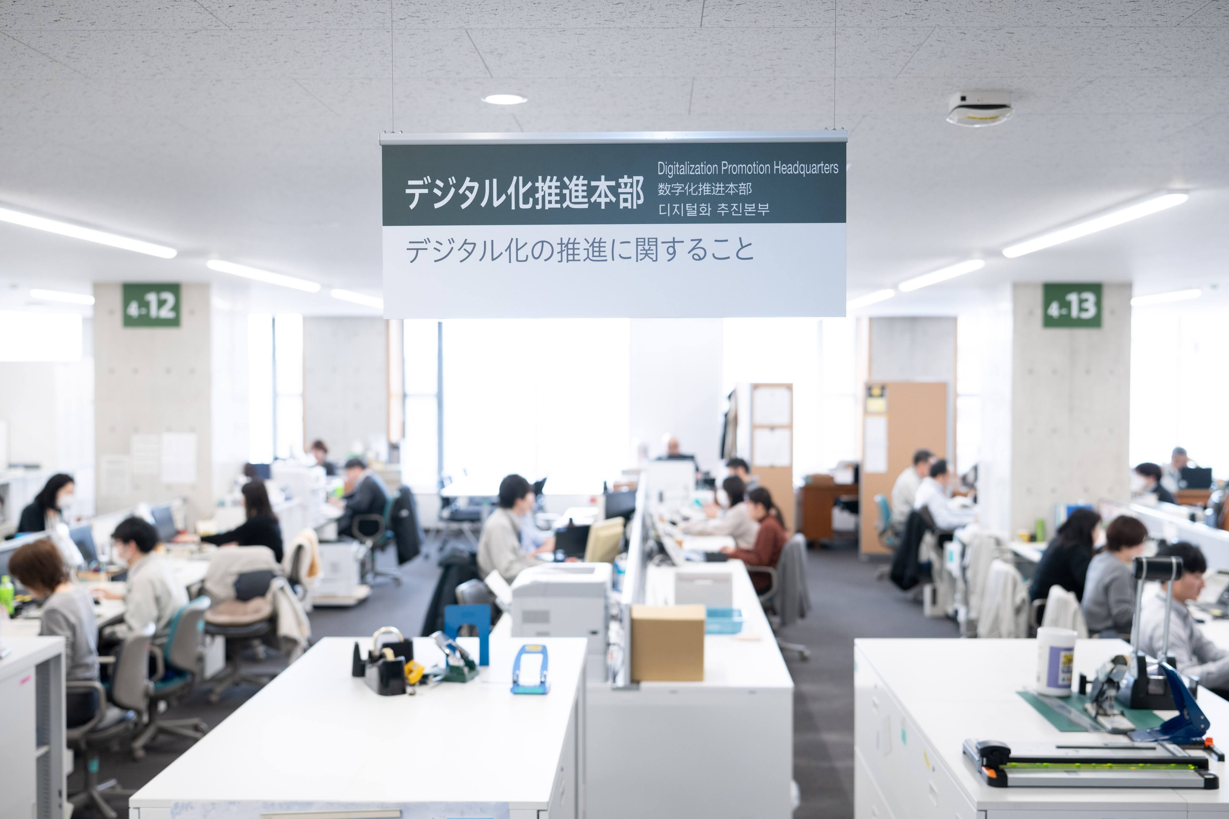 秋田市デジタル化推進本部の執務スペースを写した写真。天井から吊り下げた看板には「デジタル化推進本部 デジタル化の推進に関すること」と書かれている。執務スペースには、着席して作業する職員の姿が見える。