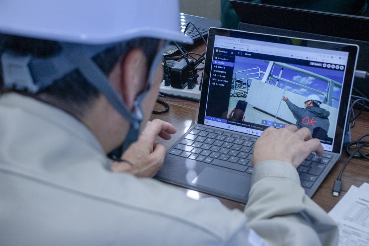 白いヘルメットを着用した担当者が、ノートパソコンの画面を操作している。画面には、ライブ映像として現場でコンクリート壁を計測している作業員の映像が映し出されている。映像には「OK」の文字が赤く書き込まれている。