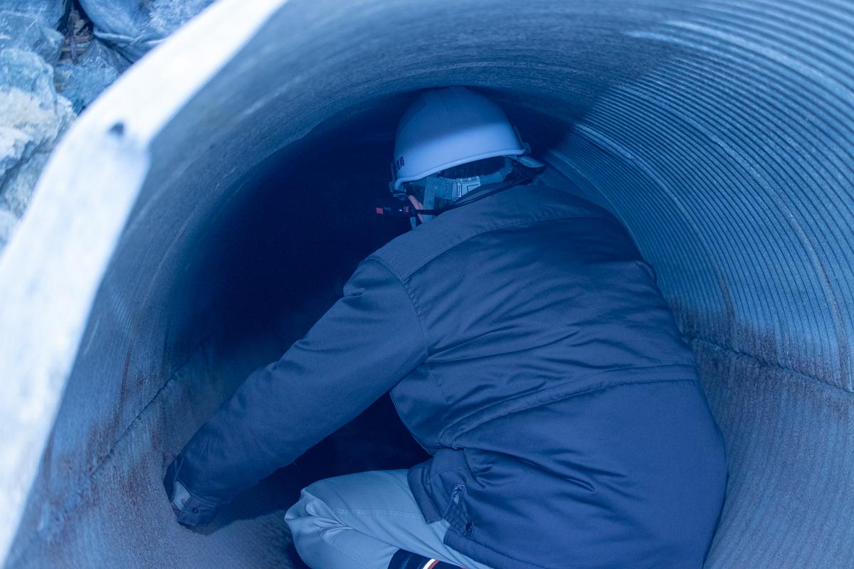 白いヘルメットをかぶった人物が、大型の円形コンクリート管（管渠）の内部に入り点検作業を行っている。ヘルメットにはカメラ機器が装着されており、内部の状況を記録している。