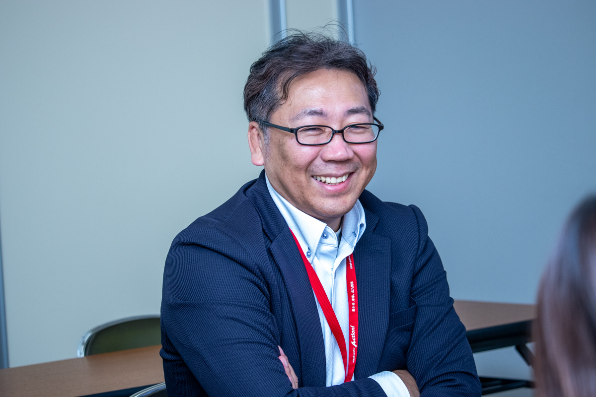 インタビューに応じる北九州市の渡辺係長。着席し腕を組みながら笑顔で話している。紺色のジャケットに白いシャツを着用し、赤いストラップの名札を首から下げている。