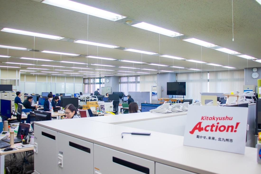 蛍光灯が並ぶ広いオープンオフィスで、複数の職員がデスクに向かって業務を行っている。手前のキャビネット上には「Kitakyushu Action! 動かせ、未来。北九州市」と書かれた白いプレートが置かれている。奥には大型モニターや仕切りパネルが設置されている。