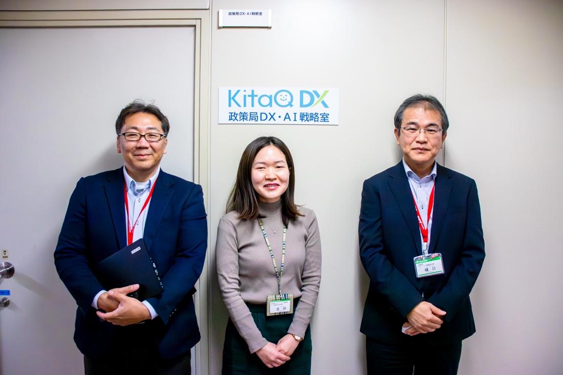 「KitaQ DX 政策局DX・AI戦略室」と書かれたロゴプレートが貼られた扉の前に3名が並んでいる。左側から渡辺係長、金田係長、樋口次長。3名とも正面を向いて微笑んでいる。