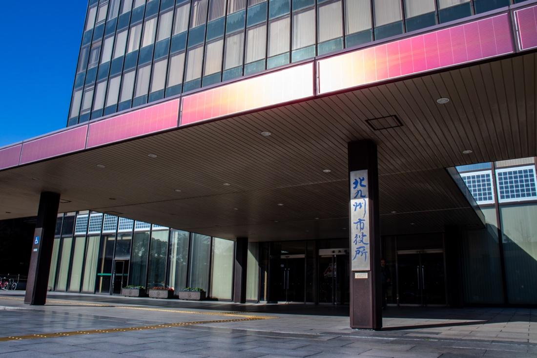 快晴の青空のもと、北九州市役所の正面玄関を撮影した写真。手前の柱には「北九州市役所」と縦書きされ、その奥に広い庇（ひさし）が張り出した玄関エントランスが見える。玄関前の広場は石畳で整備されており、左手には自転車が数台駐輪している。