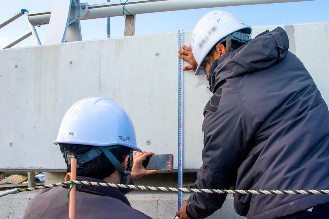 屋外の工事現場で、白いヘルメットをかぶった2名の作業員が、コンクリートの壁面に対してスケール（巻き尺）を当てて計測作業を行っている。手前の作業員はスマートフォンで撮影しており、奥の作業員がスケールを壁に押し当てている。背景には鉄骨構造物と青空が見える。ロープが手前に張られている。