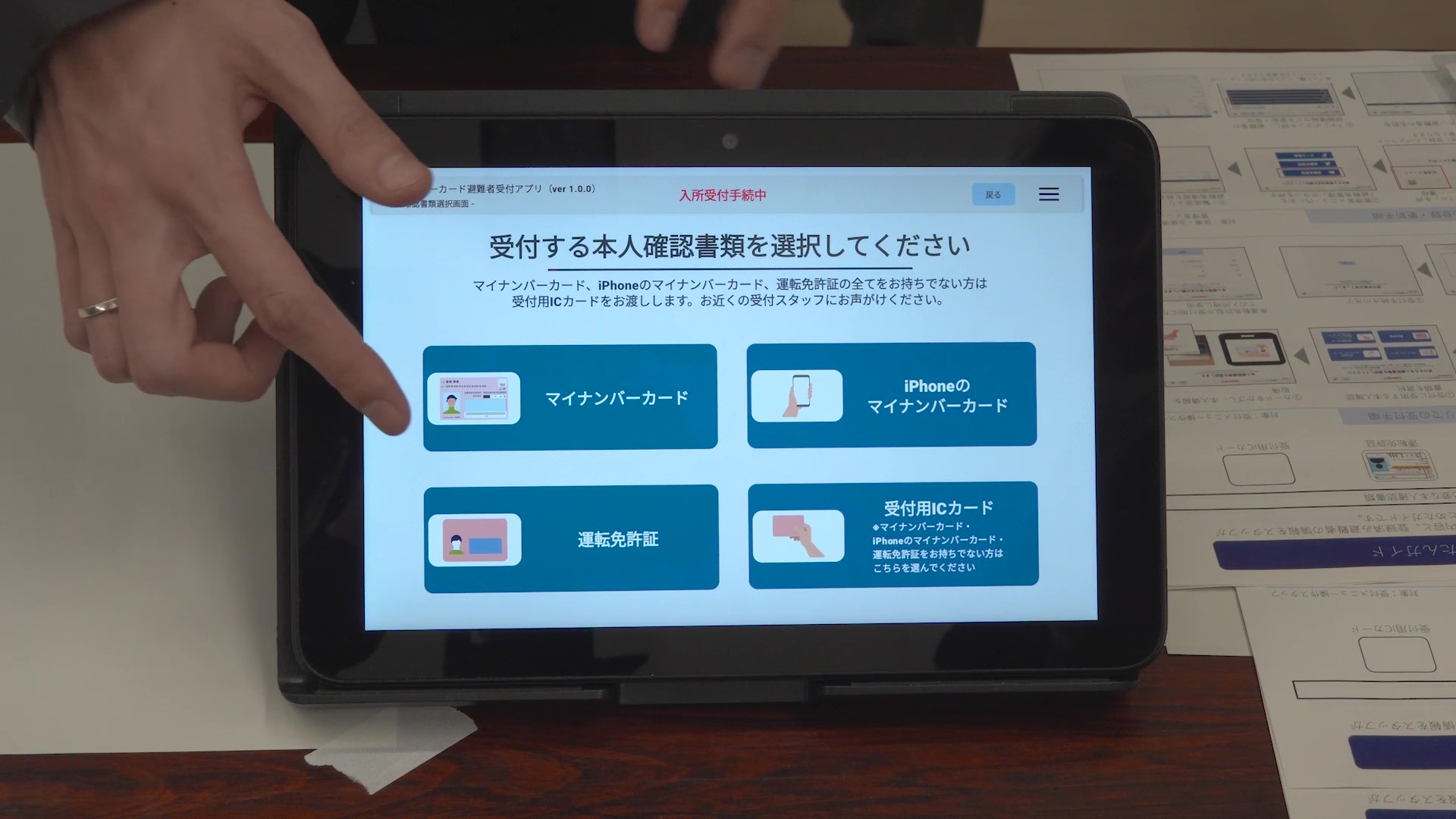 机の上に置かれたタブレット端末を、操作している写真。タブレットの画面には「マイナンバーカード遊覧者受付アプリ(ver 1.0.0)」の「本人確認書類選択画面」が表示されており、画面上部に「入所受付手続中」と赤字で示されている。画面中央には「受付する本人確認書類を選択してください」という見出しがあり、その下に「マイナンバーカード」「iPhoneのマイナンバーカード」「運転免許証」「受付用ICカード(マイナンバーカード・iPhoneのマイナンバーカード・運転免許証をお持ちでない方はこちらを選んでください)」の4つの青いボタンが2×2のグリッド状に配置されている。タブレットの背後から画面を示す指は「マイナンバーカード」ボタンを指し示している。