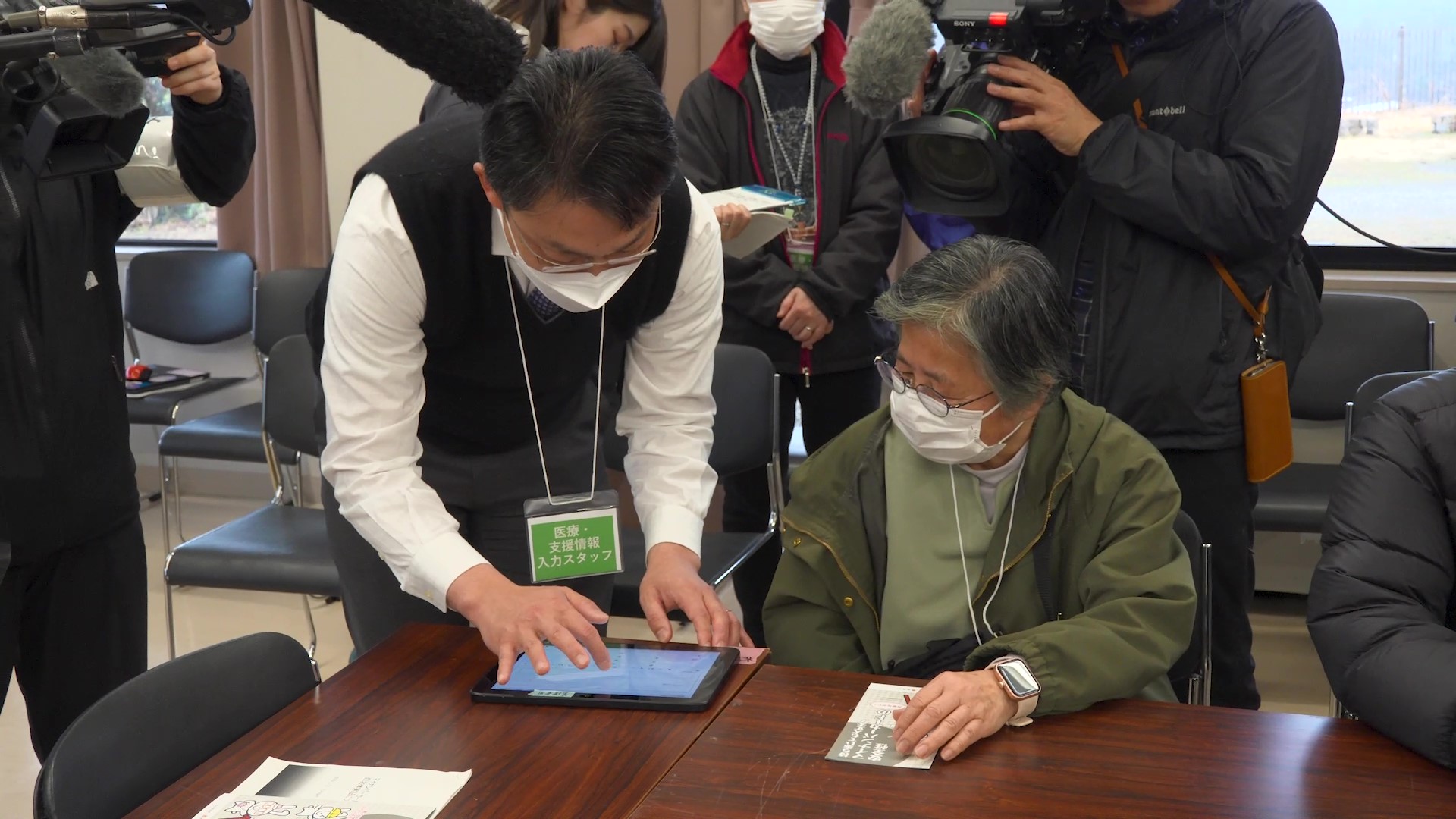 名張市での実証の様子。「医療・支援情報入力スタッフ」という名札を首から下げた人物がタブレット端末を操作し、避難者役と推認される着席した人物に聞き取りを行っている。二人の背後には、映像撮影をする人物が写っている。