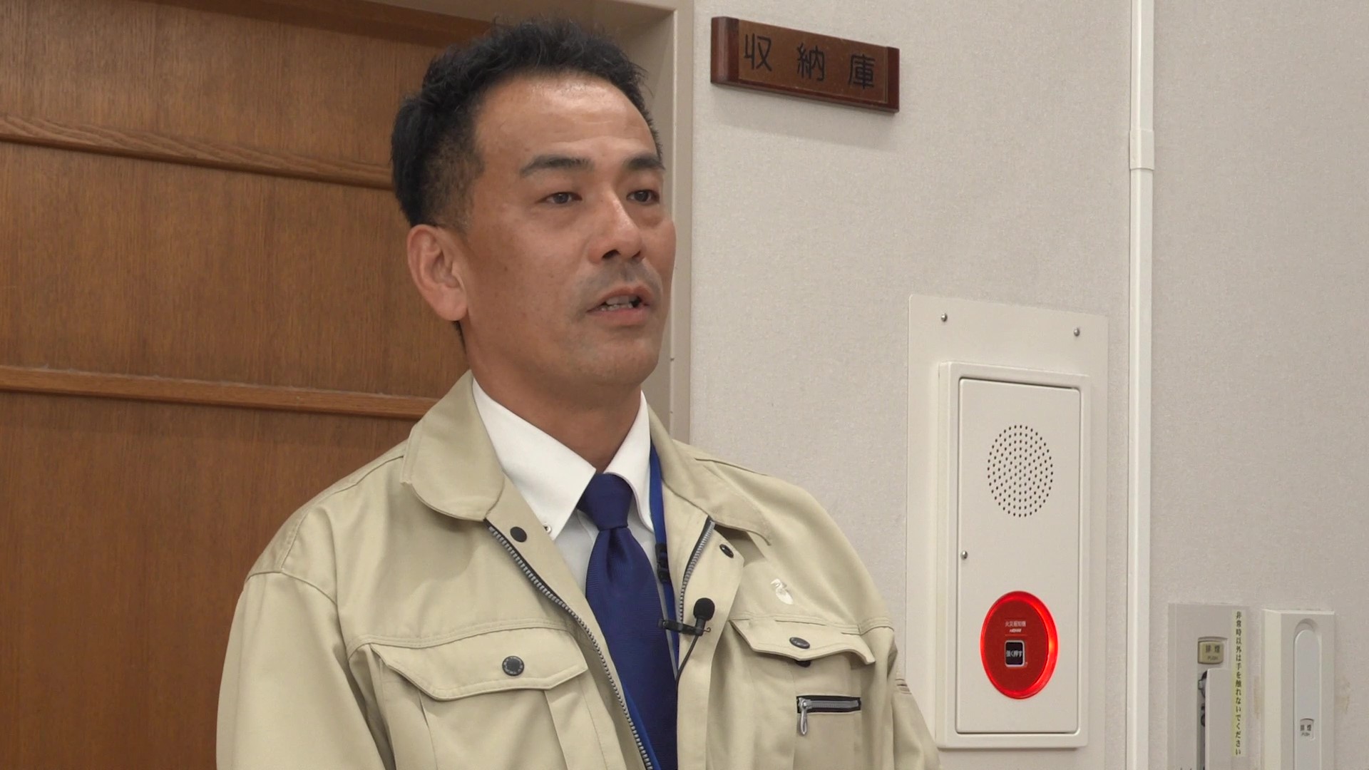 ベージュ色の作業着を着用した稲垣係長が、やや斜め前方を向いて話している。背景には木目調のパネルと白い壁があり、壁には「収納庫」と書かれたプレートが掲示されている。