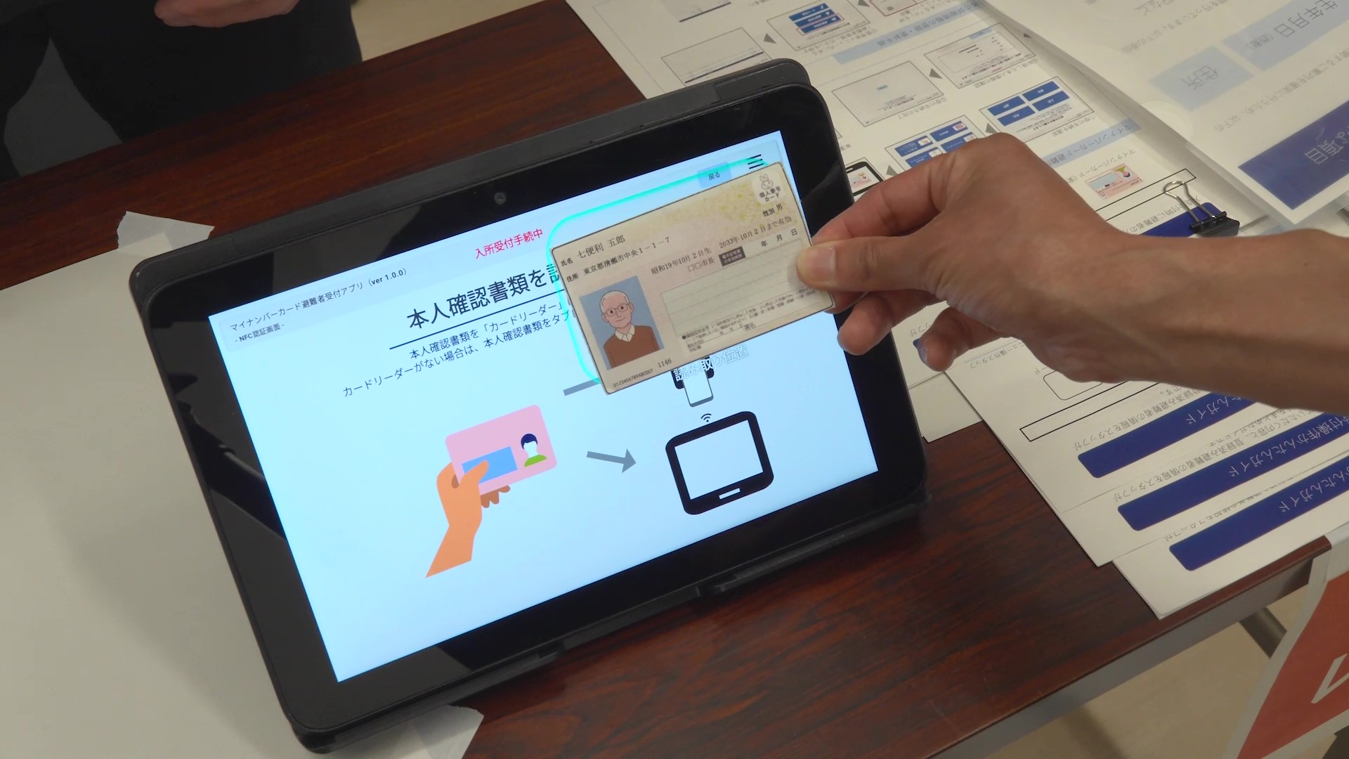 木製のテーブルの上に置かれたタブレット端末に向かって、人物の右手がマイナンバーカード(※ダミーカード)をかざしている場面の写真。タブレット画面には『マイナンバーカード遮難者受付アプリ(ver 1.0.0)』と表示されており、『本人確認書類を読み取り中』という状態を示している。画面中央には『本人確認書類をカードリーダーにかざしてください』という案内文と、カードをカードリーダーにかざす操作手順を示したイラスト(ピンク色のカードを持つ手のイラストと、矢印、黒いカードリーダーのイラスト)が表示されている。