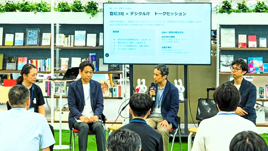 左から、デジタル庁の小田部巧参事官補佐、明治安田生命保険相互会社の小谷修一氏、株式会社セブン銀行の三浦龍矢氏、マネックス証券株式会社の法貴大輔氏が椅子に座って、トークセッションをしている様子。4者の後ろにはスクリーンがあり、「登壇3社×デジタル庁 トークセッション」の文字が見える。写真には4者の話を聞く参加者の後ろ姿が5人ほど確認できる。