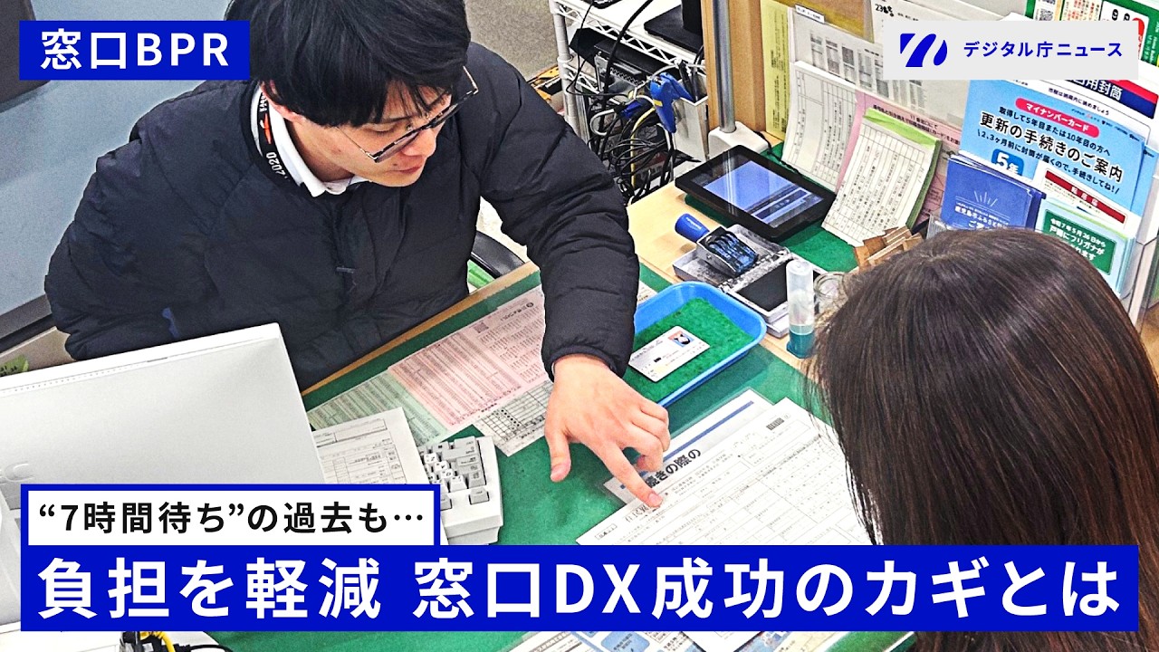 デジタル庁ニュースの動画サムネイル画像。左上に「窓口BPR」、右上に「デジタル庁ニュース」のロゴが表示されている。中央には、眼鏡をかけた黒いジャケット姿の男性職員が、カウンター越しに女性来庁者へ書類を指差しながら説明している場面が写っている。カウンター上にはパソコン、タブレット、各種書類が置かれており、背景にはマイナンバーカード更新手続きに関する案内ポスターが見える。画像下部には「"7時間待ち"の過去も…」というサブテキストと、「負担を軽減 窓口DX成功のカギとは」という白抜き太字のメインタイトルが青い帯の上に表示されている。